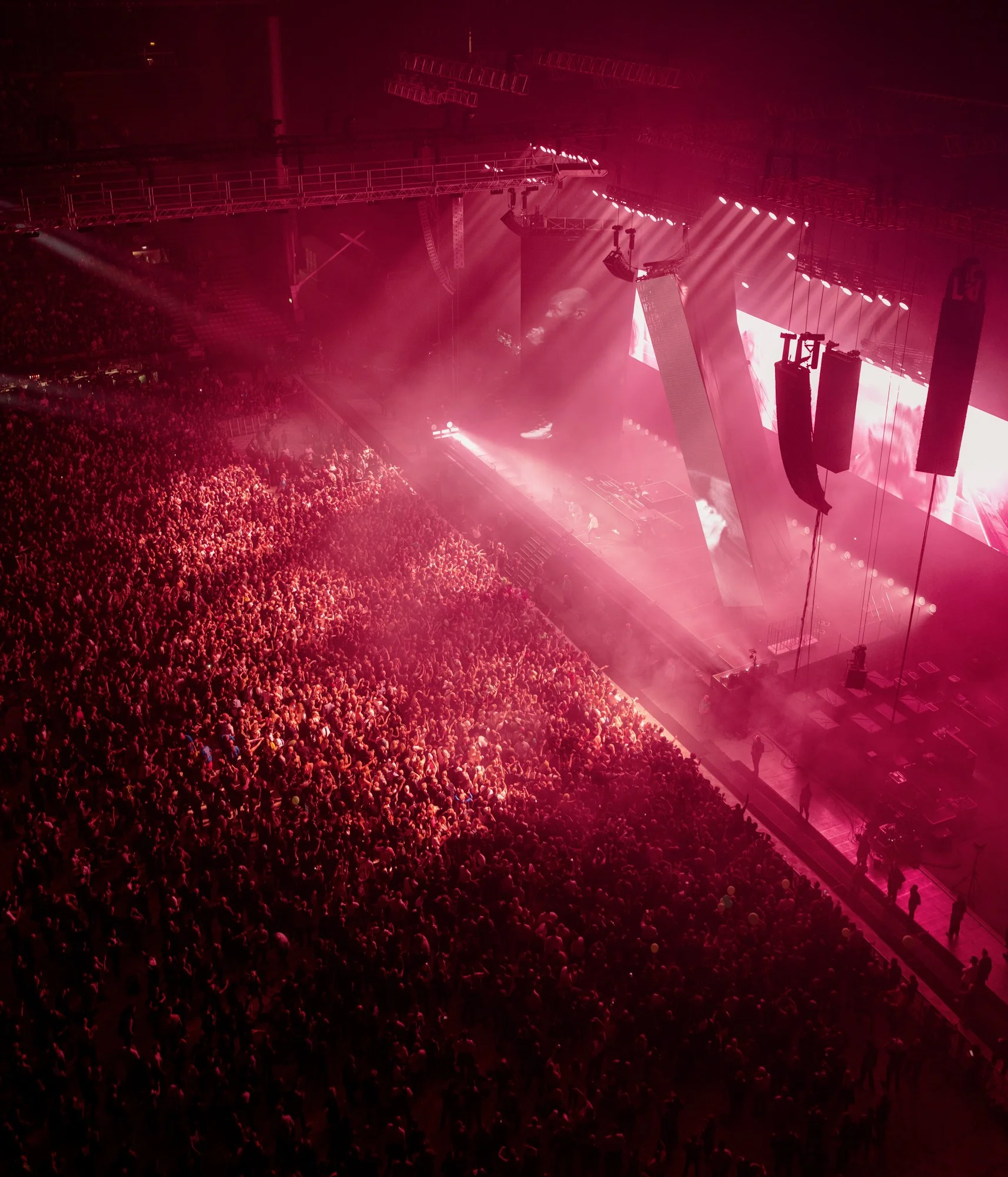 A large crowd at a concert with bright stage lights and hanging speakers.