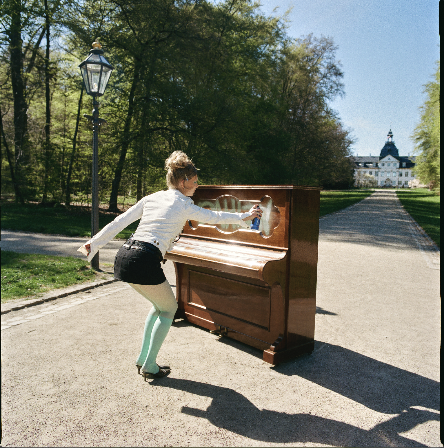 A woman in a white blouse and a black skirt is spraying a piano with a spray bottle in a park on a sunny day.
