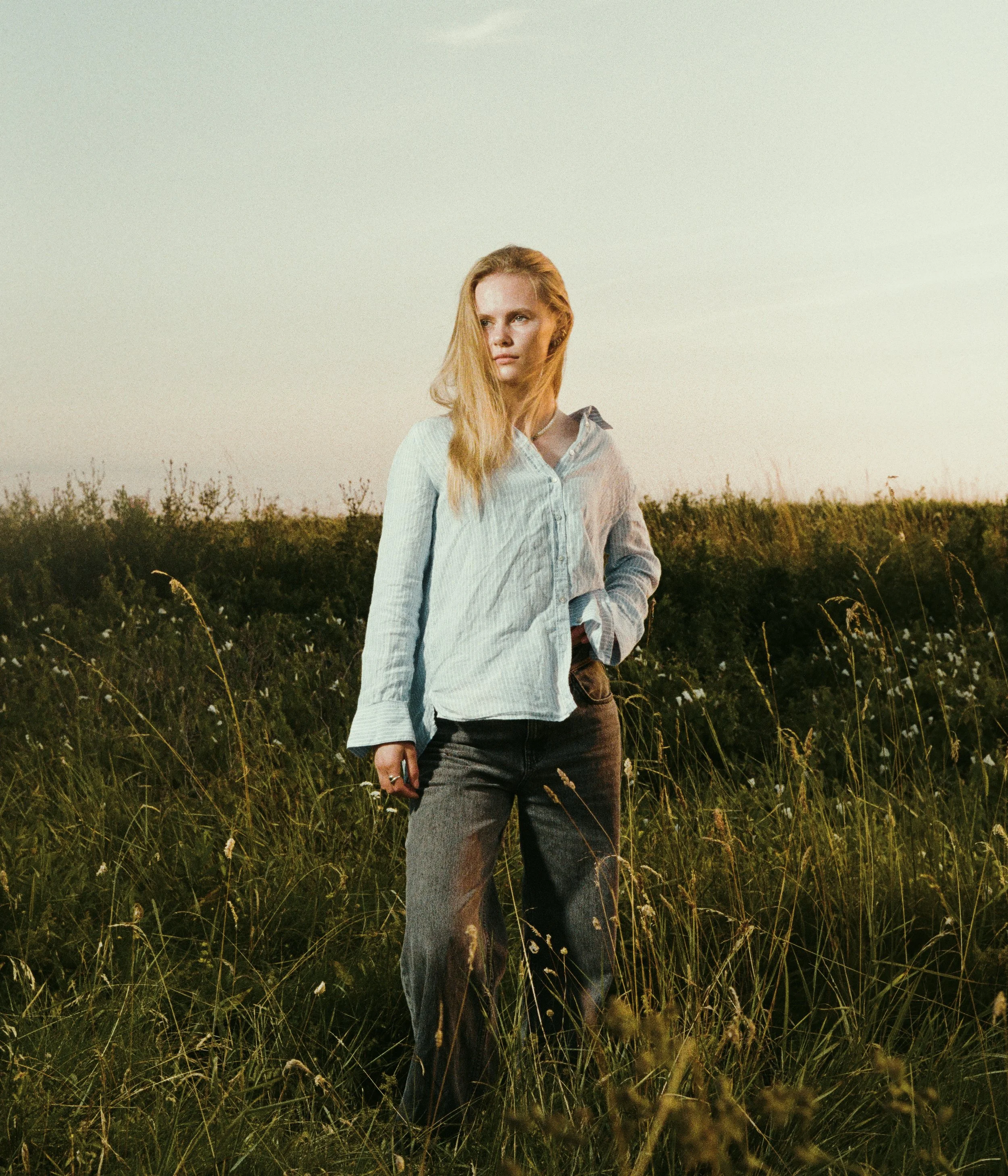 A woman with long blonde hair standing in a grassy field during sunset, wearing a light blue shirt and black jeans.
