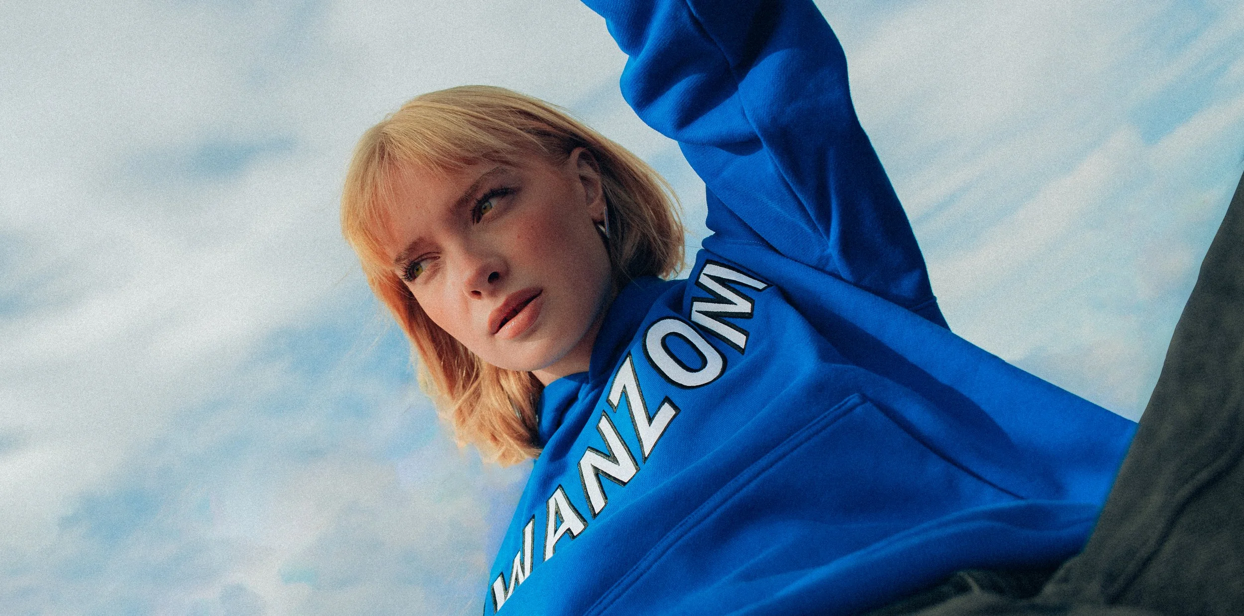A young woman with blonde hair, wearing a blue jacket with the word 'AMAZON' on it, looking into the distance against a sky with clouds.