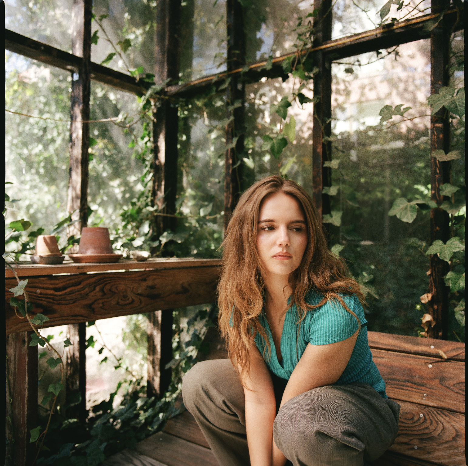 A woman with long wavy hair in a green blouse, sitting on wooden steps inside a greenhouse or garden with many green vines and plants.