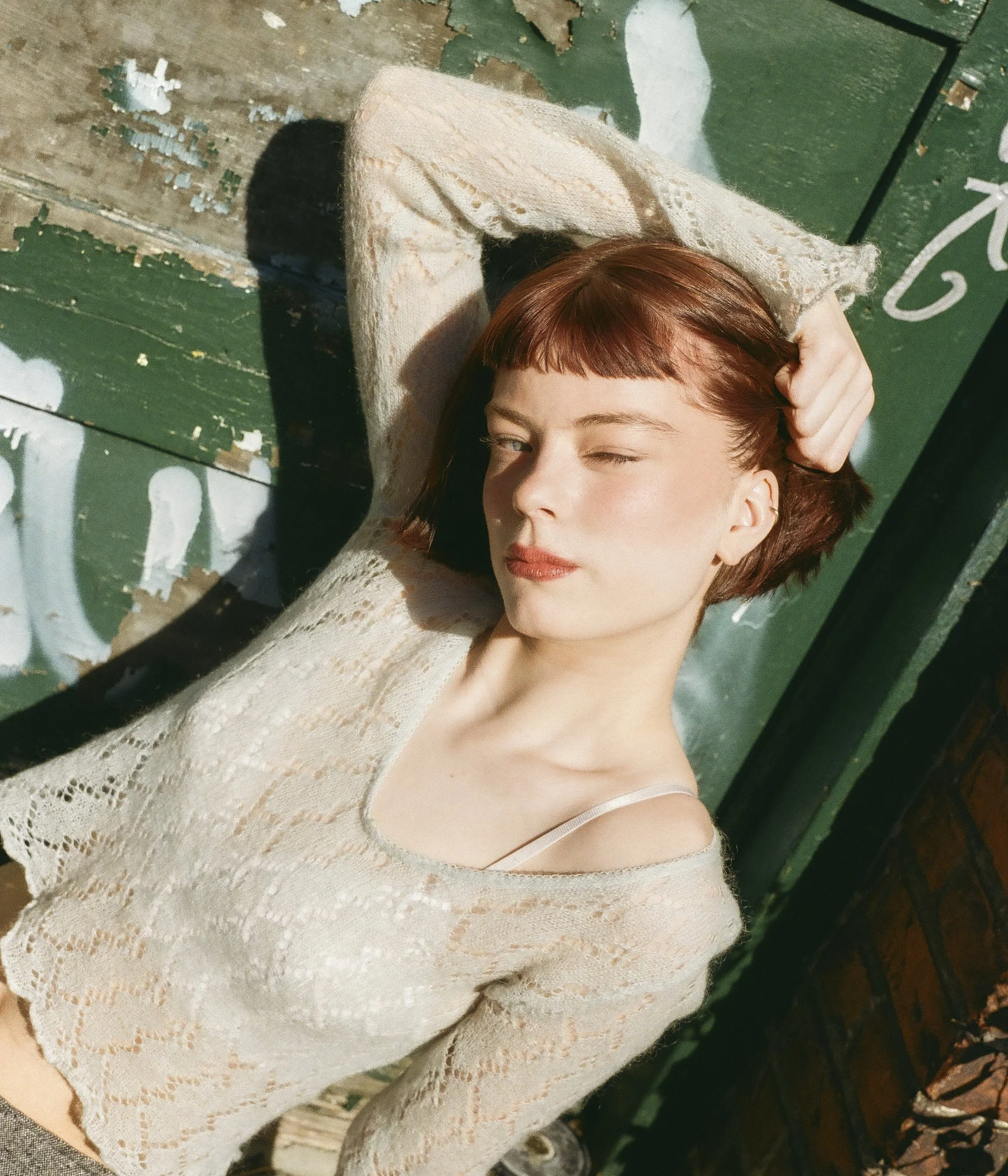 A woman with reddish-brown hair and light skin lying on a graffiti-covered green wooden surface, wearing a light, lacy sweater and a camisole, looking at the camera with one eye closed.