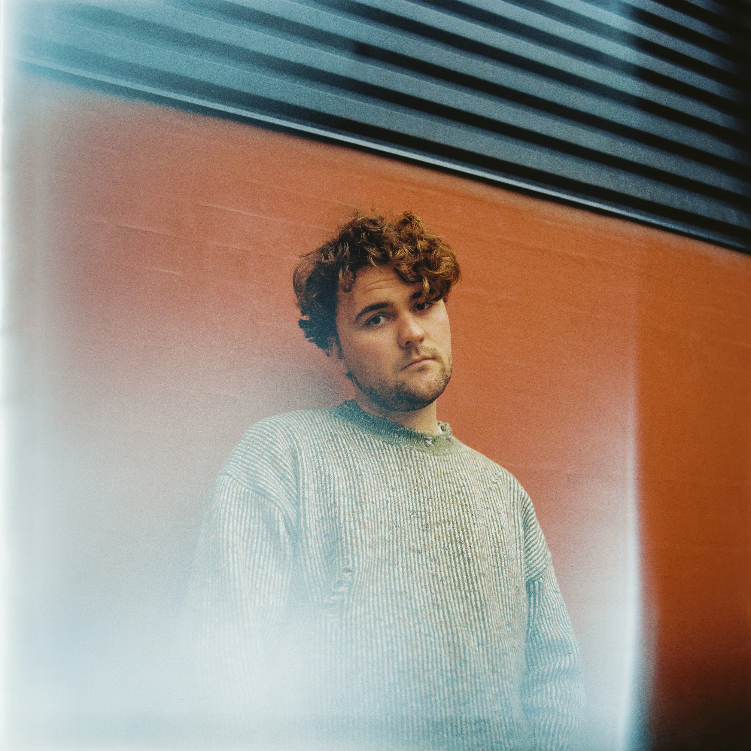 A young man with curly brown hair and a beard standing against a wall with orange and dark gray horizontal panels, wearing a gray and white striped sweater.