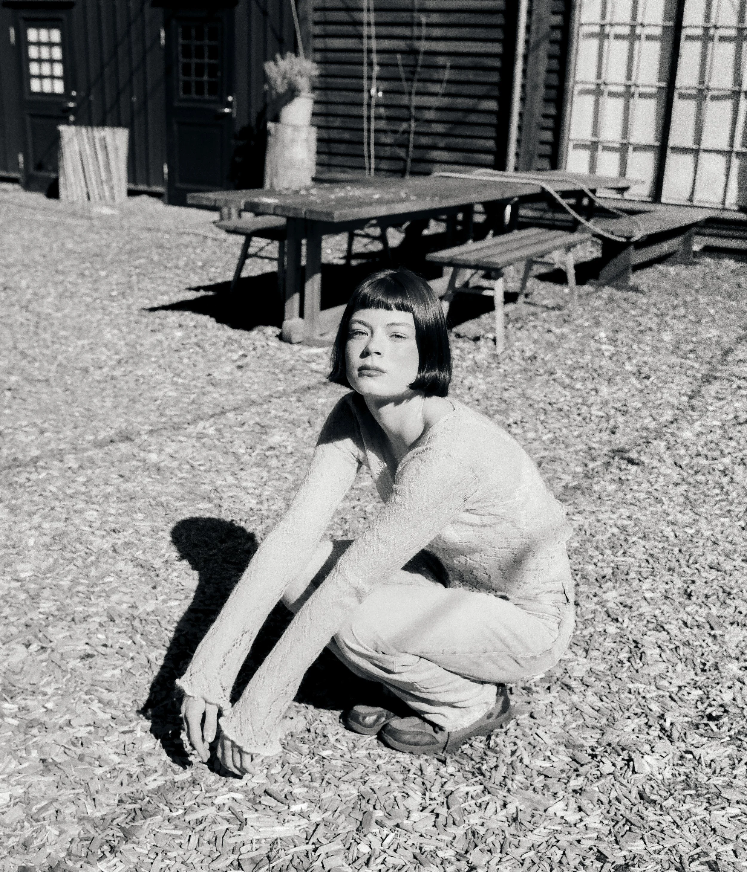 A woman with short dark hair squats on a gravel surface, looking at the camera, with a wooden outdoor table and benches, a potted plant, and a wooden fence in the background.