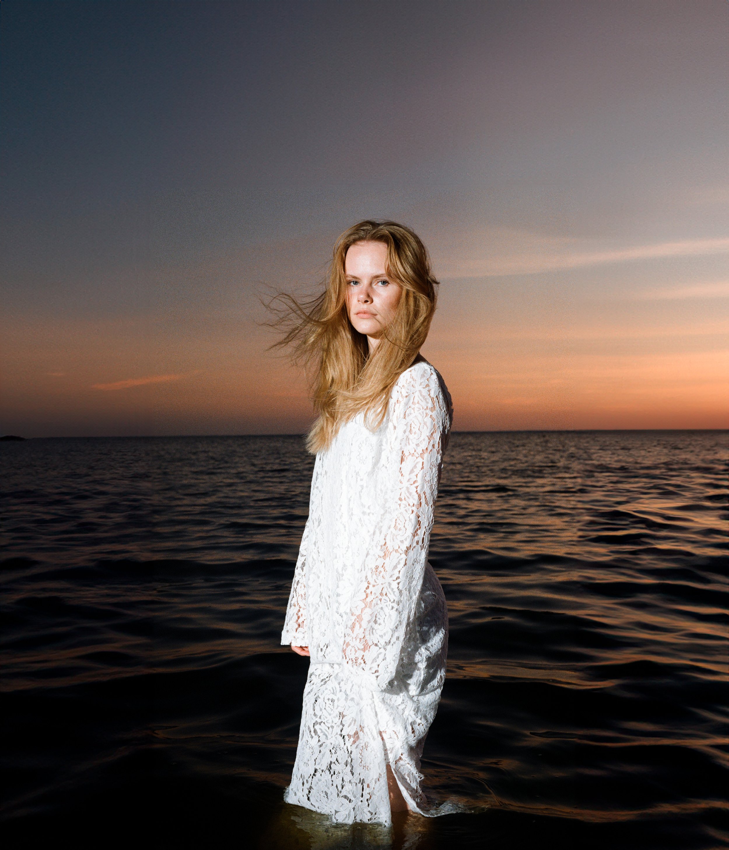 A woman in a white lace dress standing in the ocean during sunset.