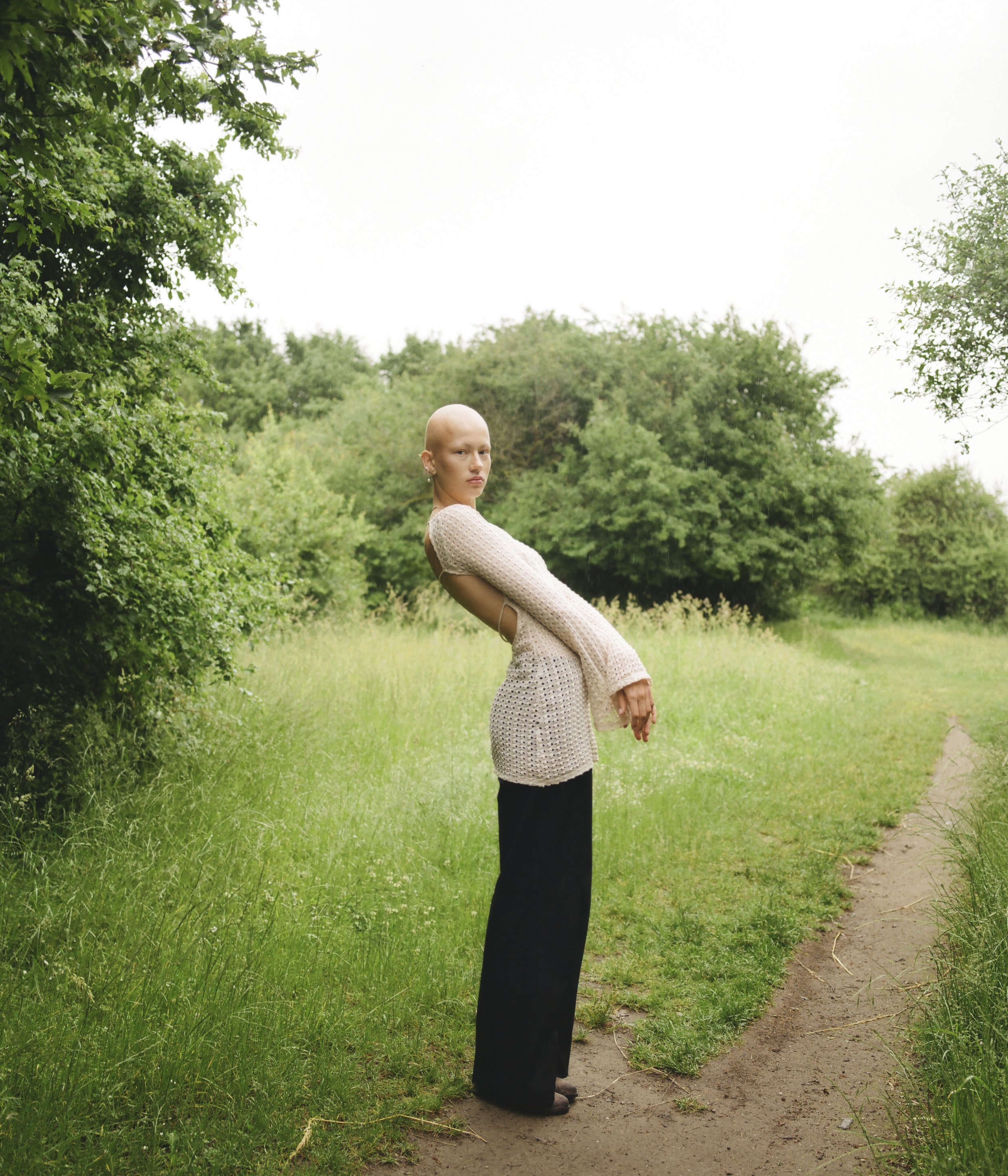 A bald woman standing on a dirt path in a green outdoor setting with trees and grass, wearing a beige crocheted top and black pants.