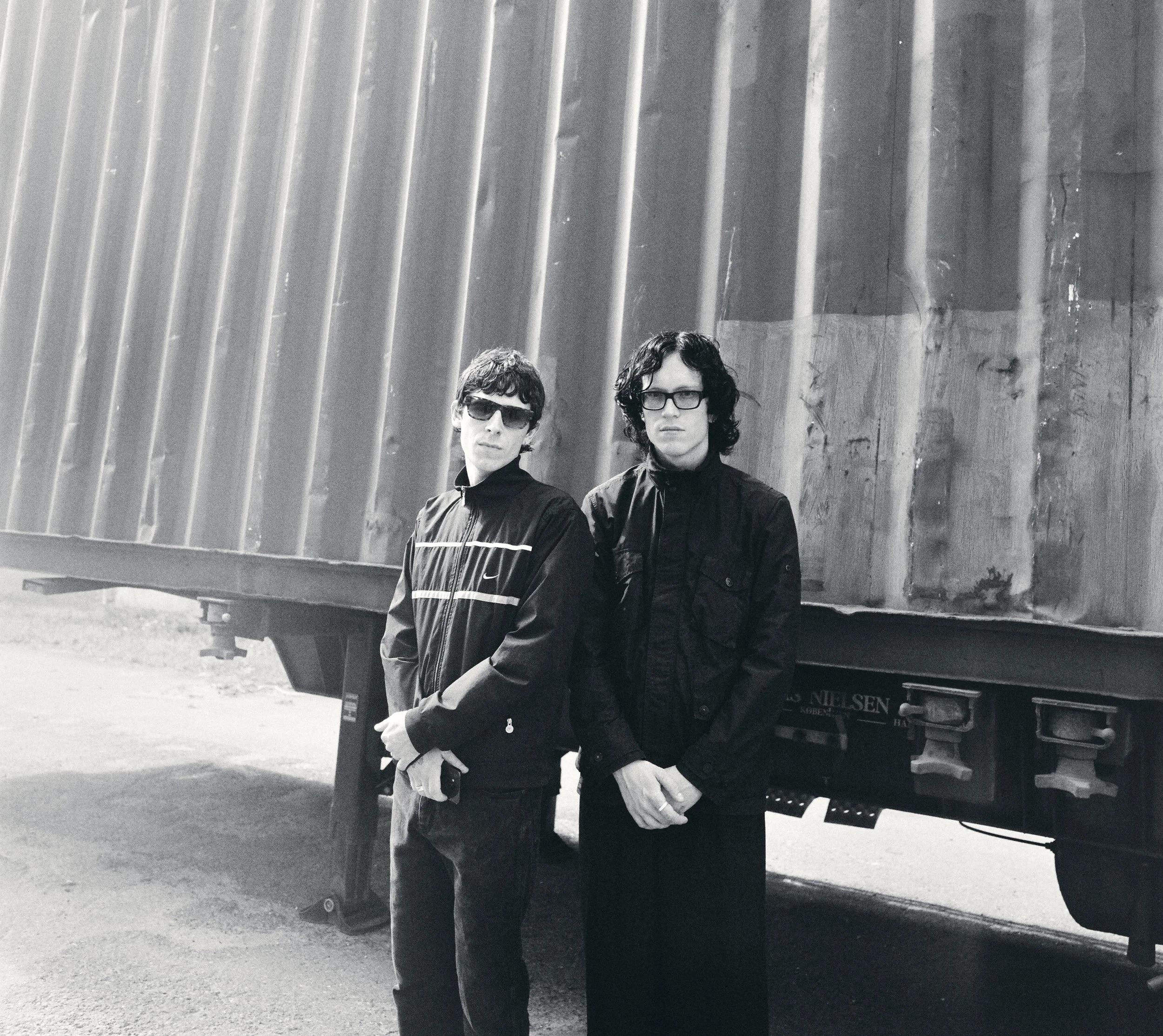 Two men with dark curly hair and glasses standing in front of a large truck with a wooden cargo container, in black and white.
