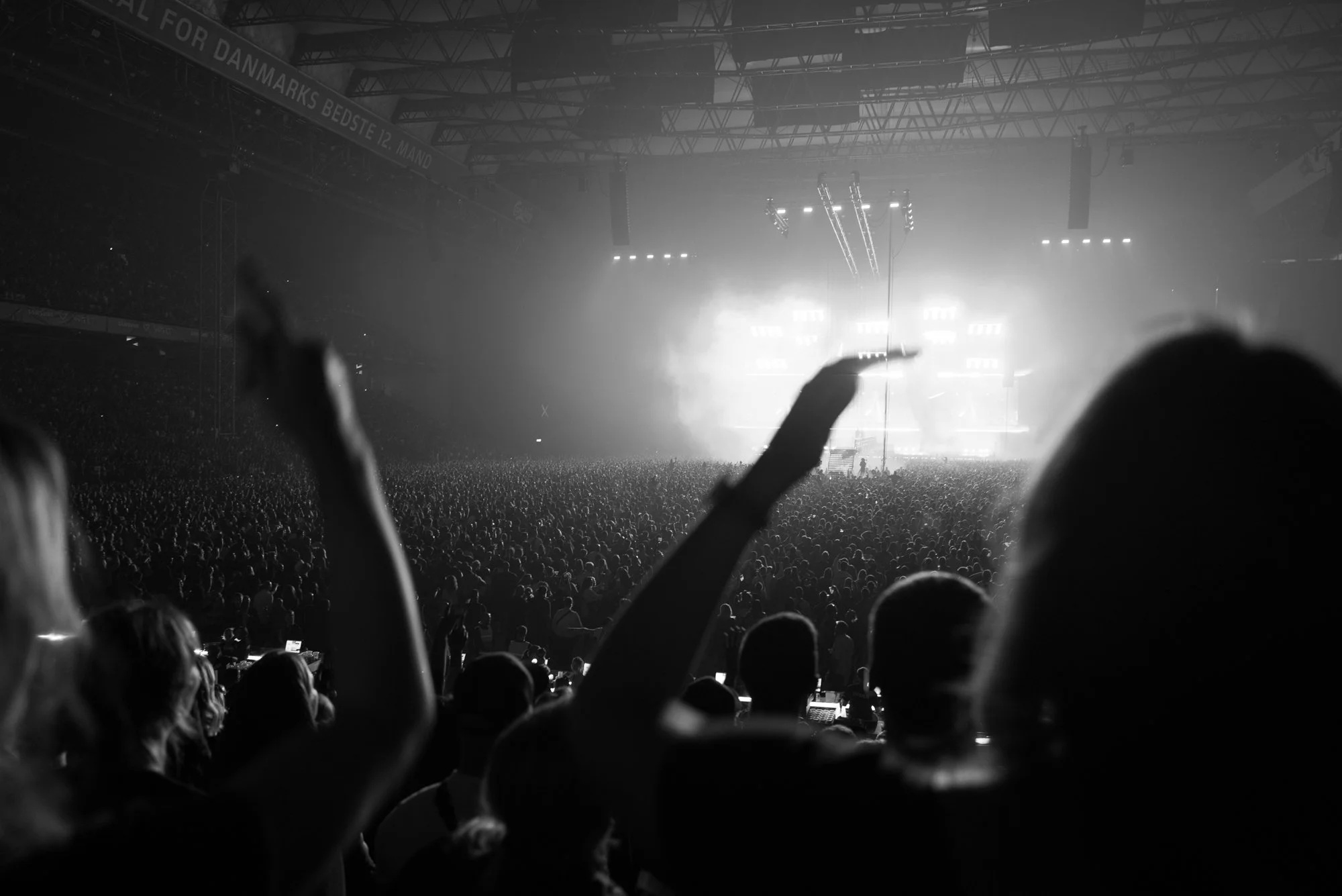 Black and white photo of a concert crowd inside a large indoor stadium, with stage lights and fog in the background, and audience members raising their hands.