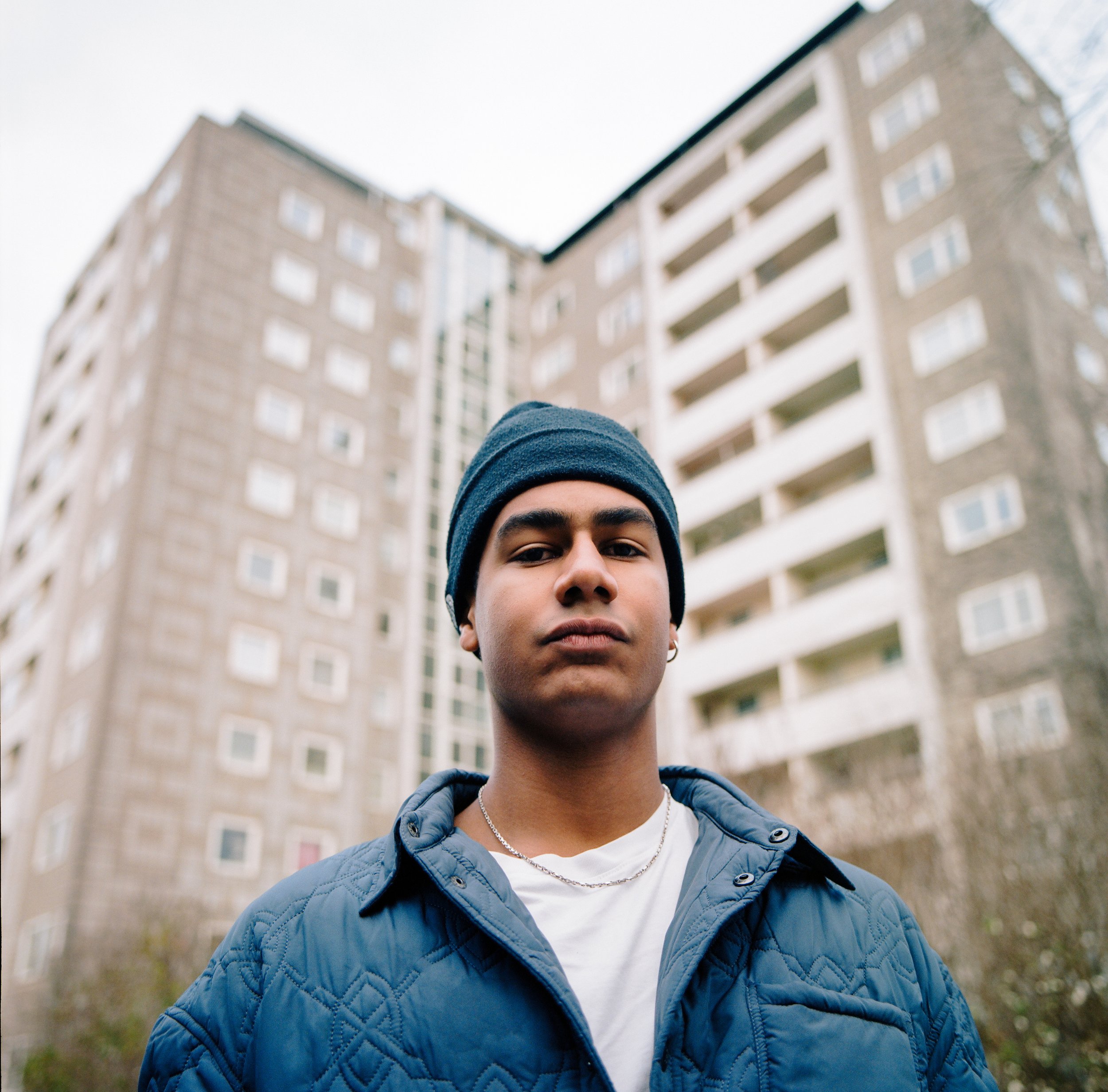 Young man in a dark beanie, blue jacket, and jewelry standing outdoors in front of a tall apartment building.
