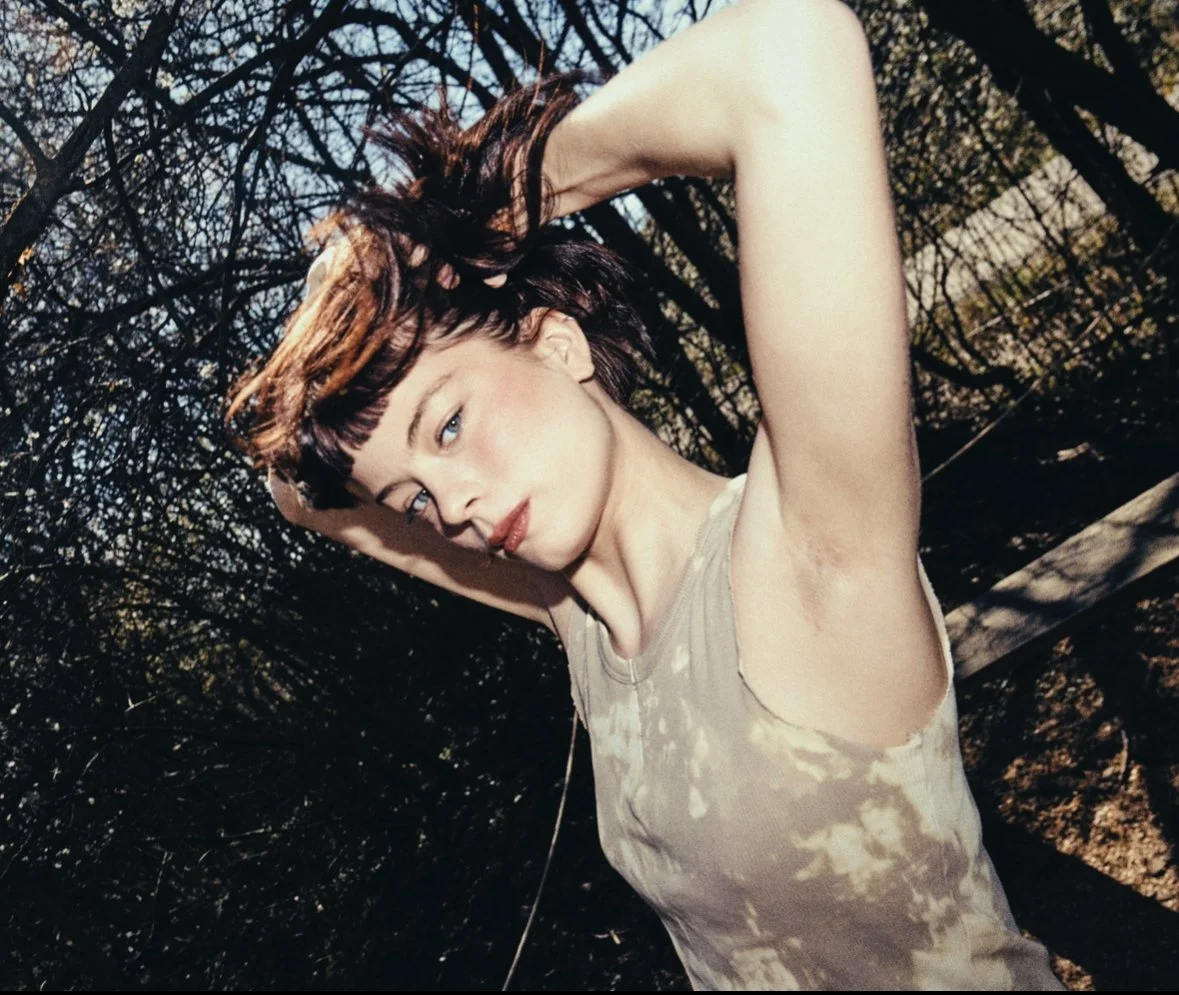 A young woman with short red hair and blue eyes, posing outdoors in a wooded area with bare branches and trees in the background. She is wearing a sleeveless beige top and has her arms raised with her hands behind her head, looking at the camera with