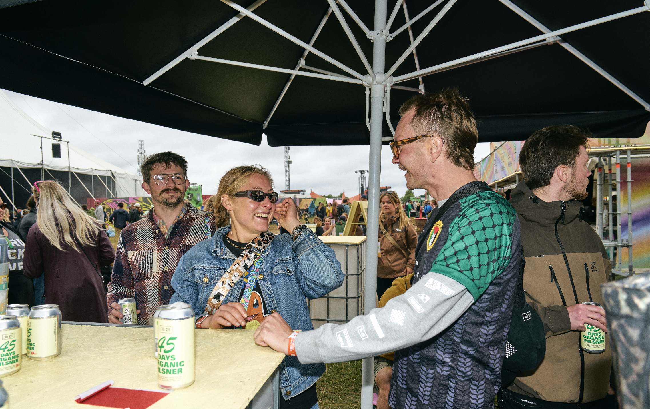 People socializing at an outdoor festival, standing around a bar with cans of organic pilsner beer.