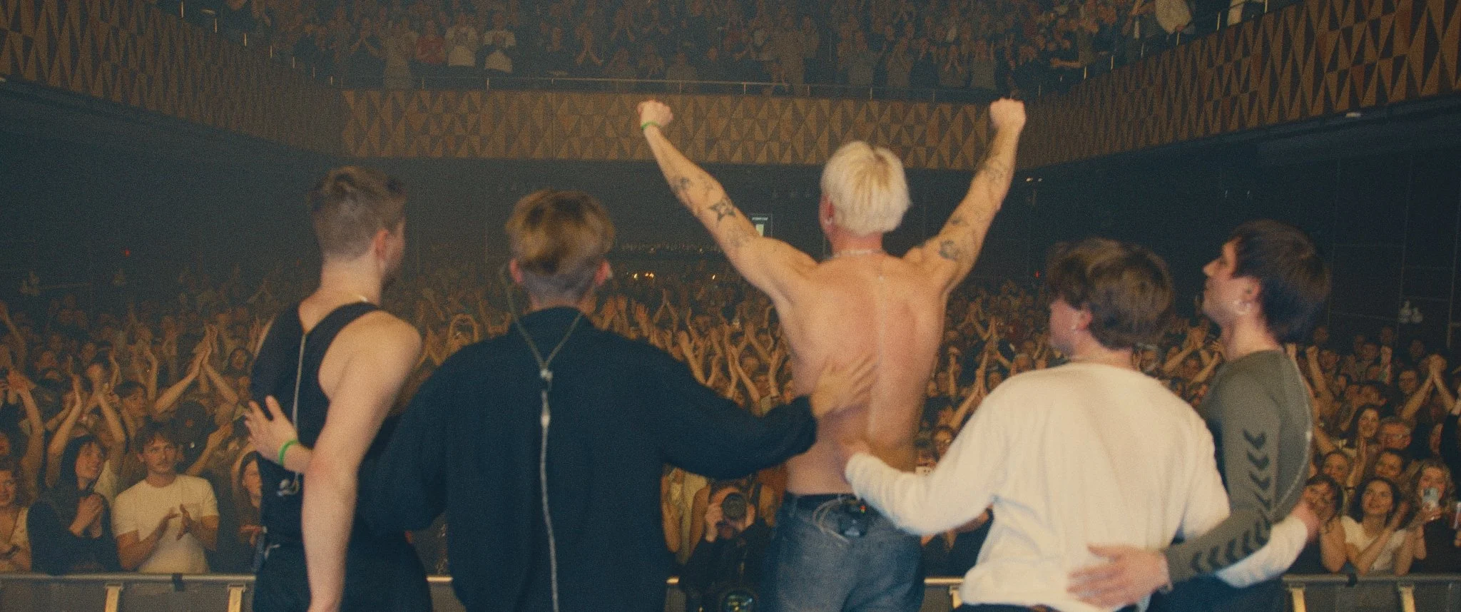 A group of young fans at a concert supporting a shirtless performer with platinum blonde hair, who has tattoos and his arms raised in victory or celebration. The fans are embracing him, and the large crowd is cheering in the background.