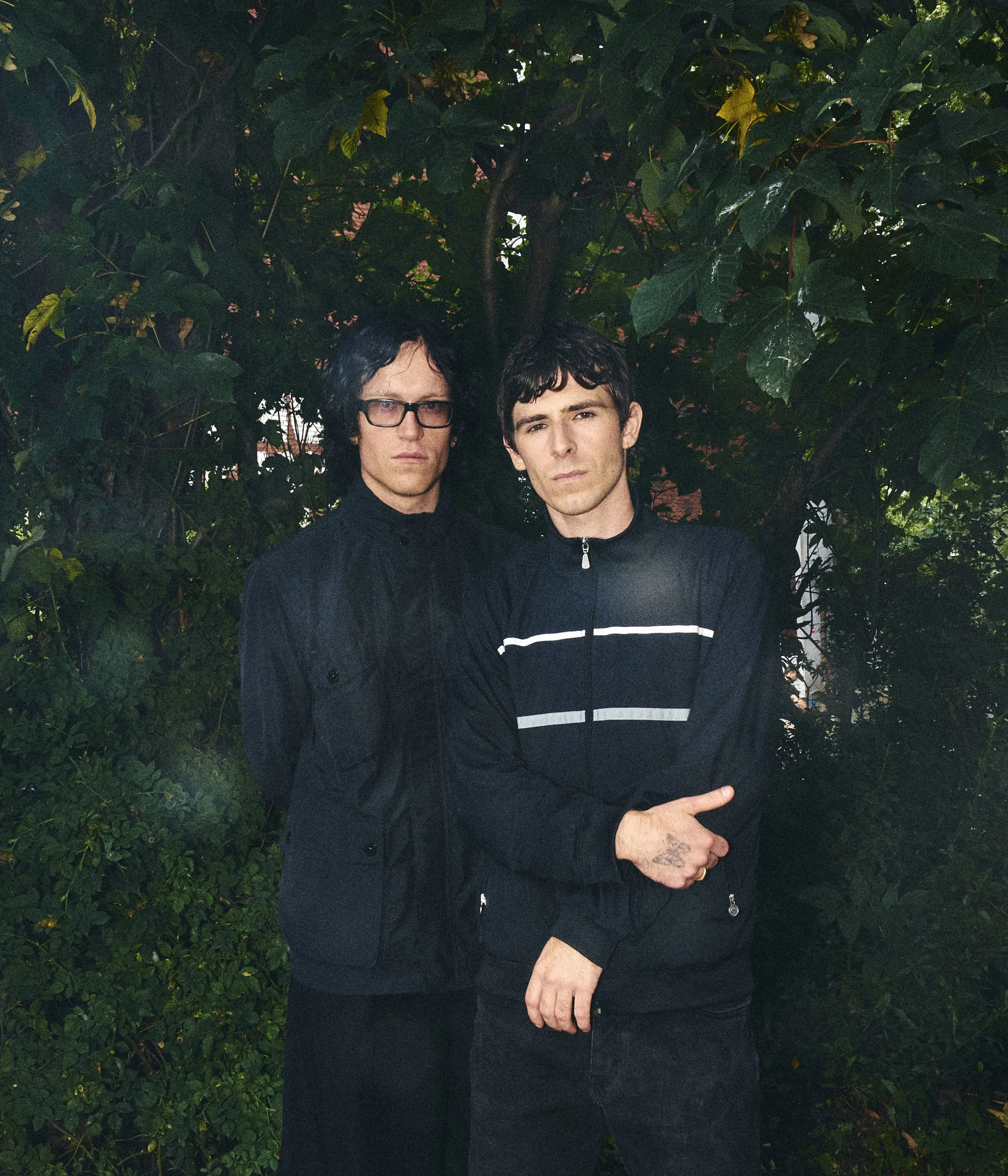Two young men standing in front of a leafy green background, dressed in dark clothing with serious expressions.