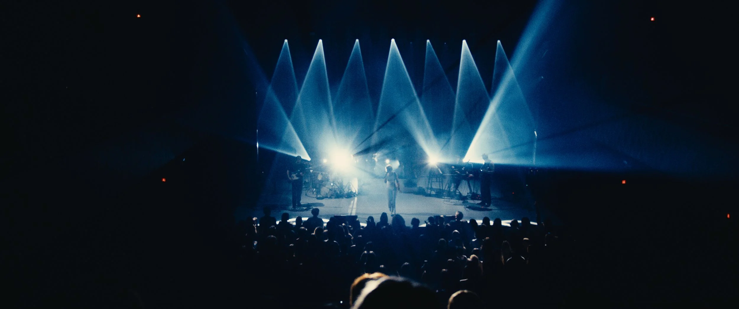 A concert stage with bright white and blue lights shining down on performers and musical equipment, with a dark audience in the foreground.