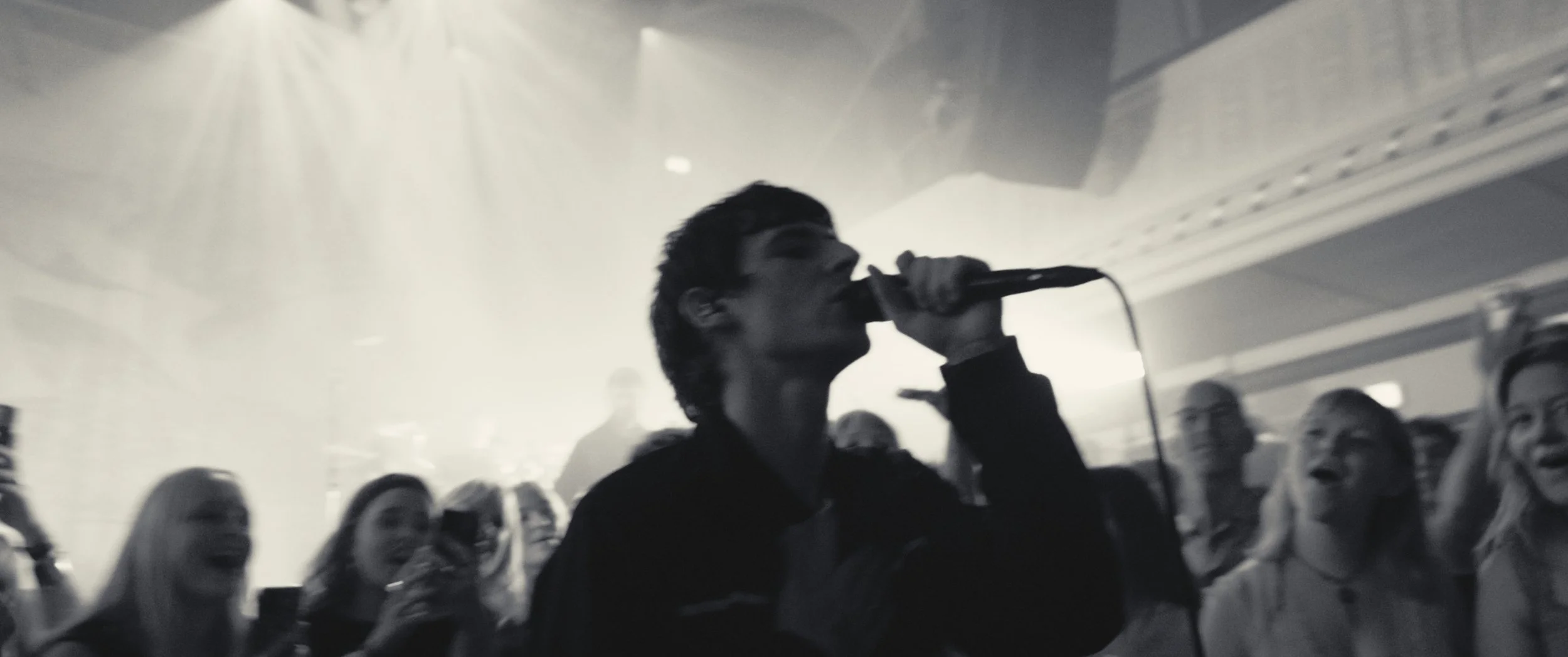 A young man singing into a microphone at a concert, surrounded by an audience with some people smiling and cheering.