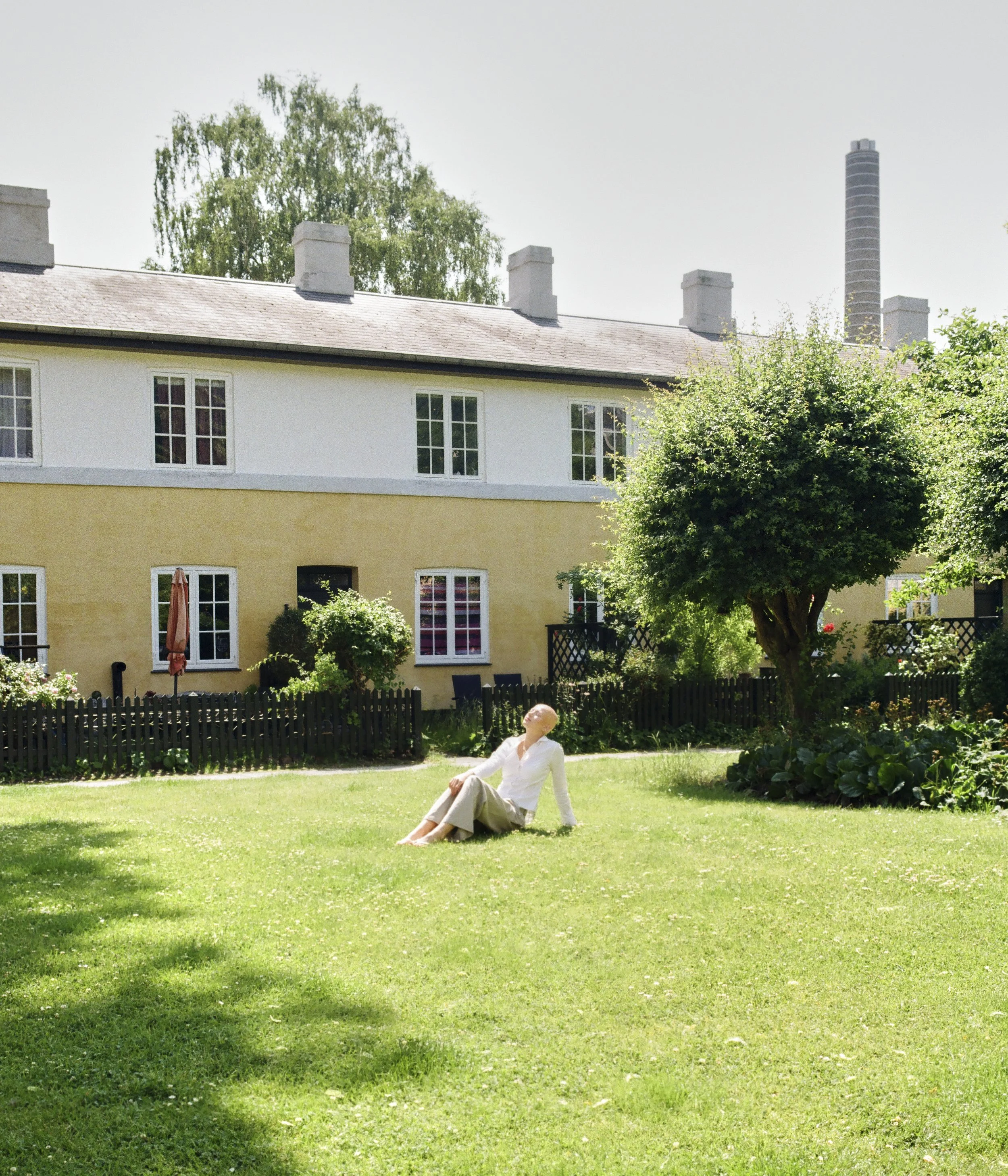A woman sitting on a lush green lawn in front of a yellow and white house with multiple windows and a black fence, surrounded by trees and garden plants on a sunny day.