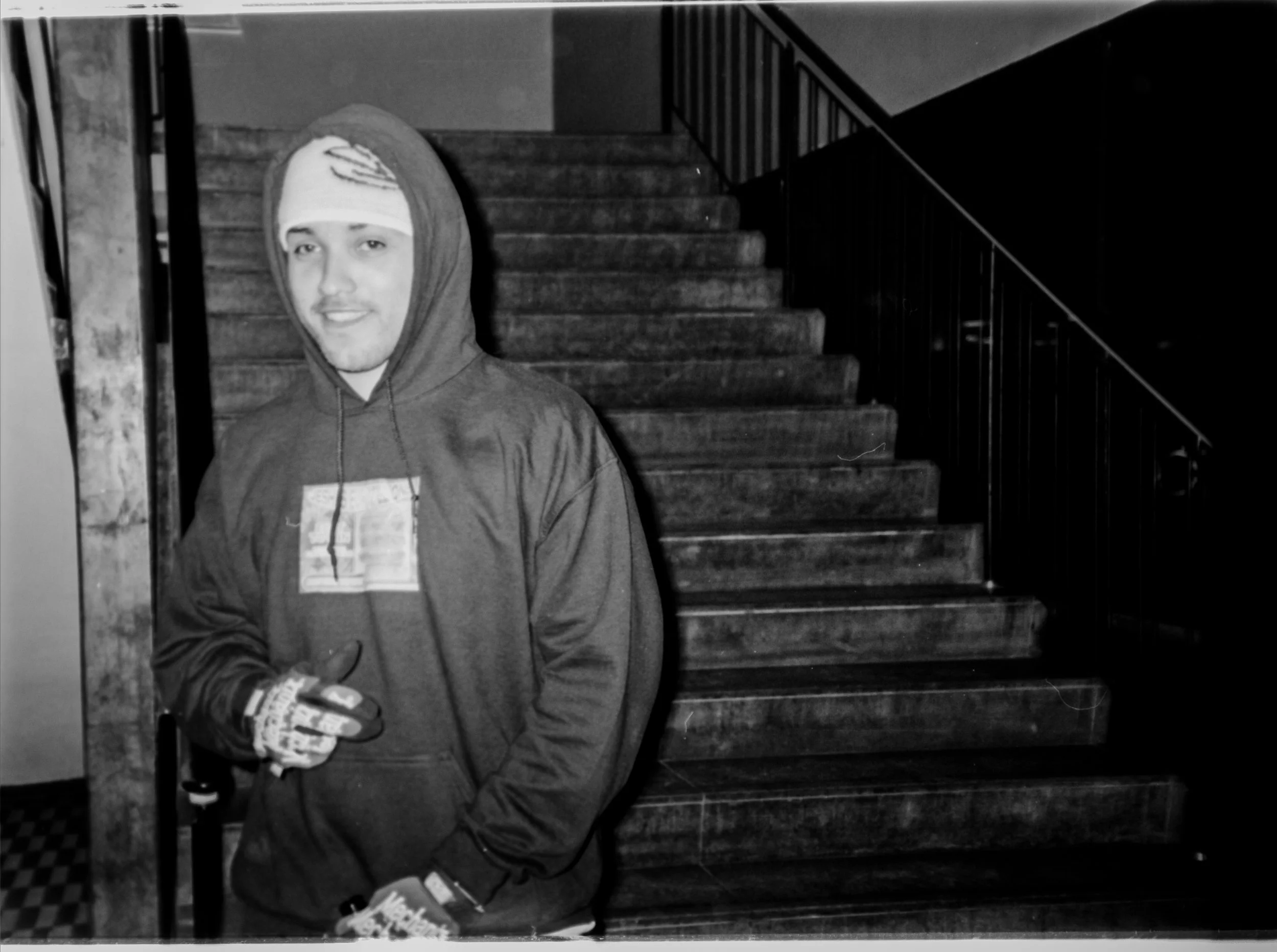 A young man in a hoodie and gloves standing indoors near a staircase, smiling, in black and white.