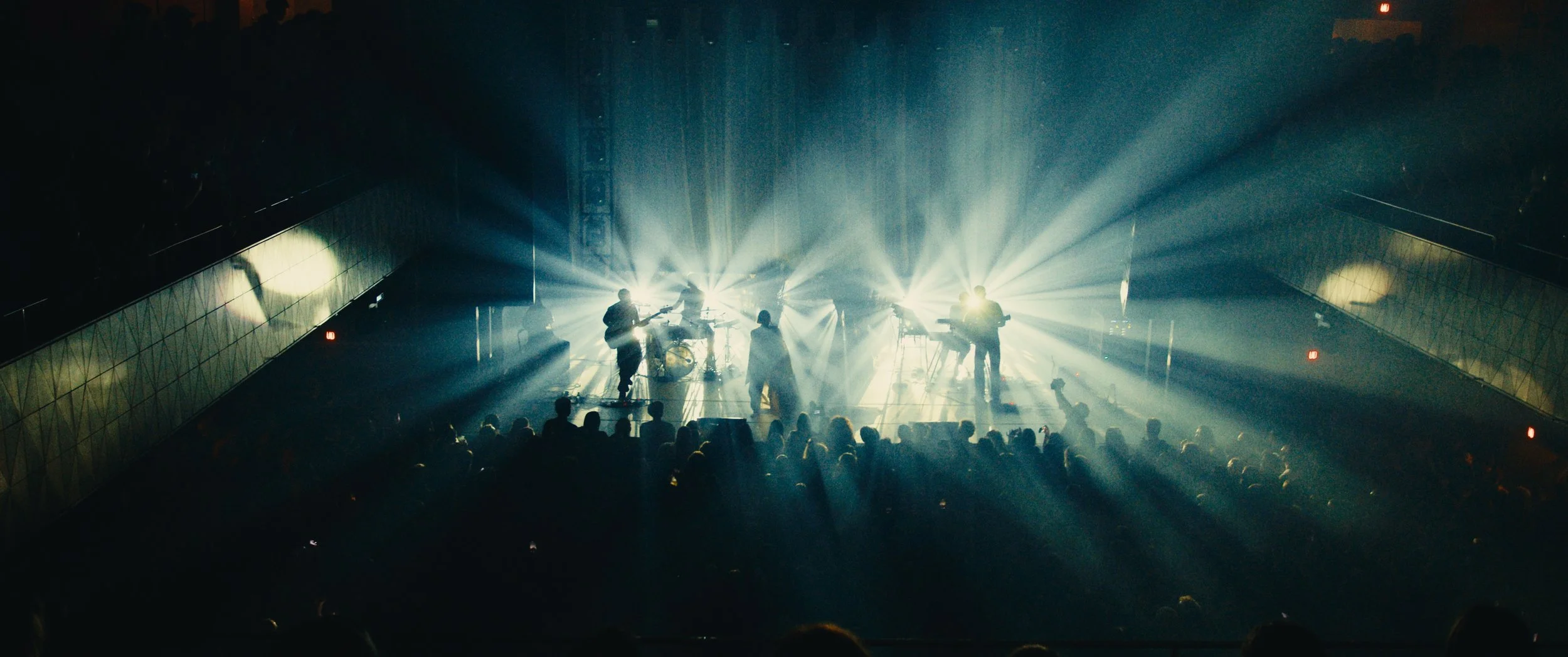 A band performing on stage with bright lights shining behind them, audience members watching from below.