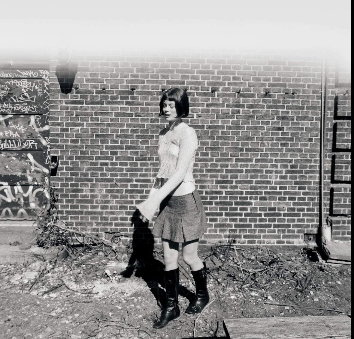 A woman with a bob haircut wearing a sweater, a pleated skirt, and tall boots stands outdoors in front of a brick wall, with graffiti partially visible on the left side of the image.