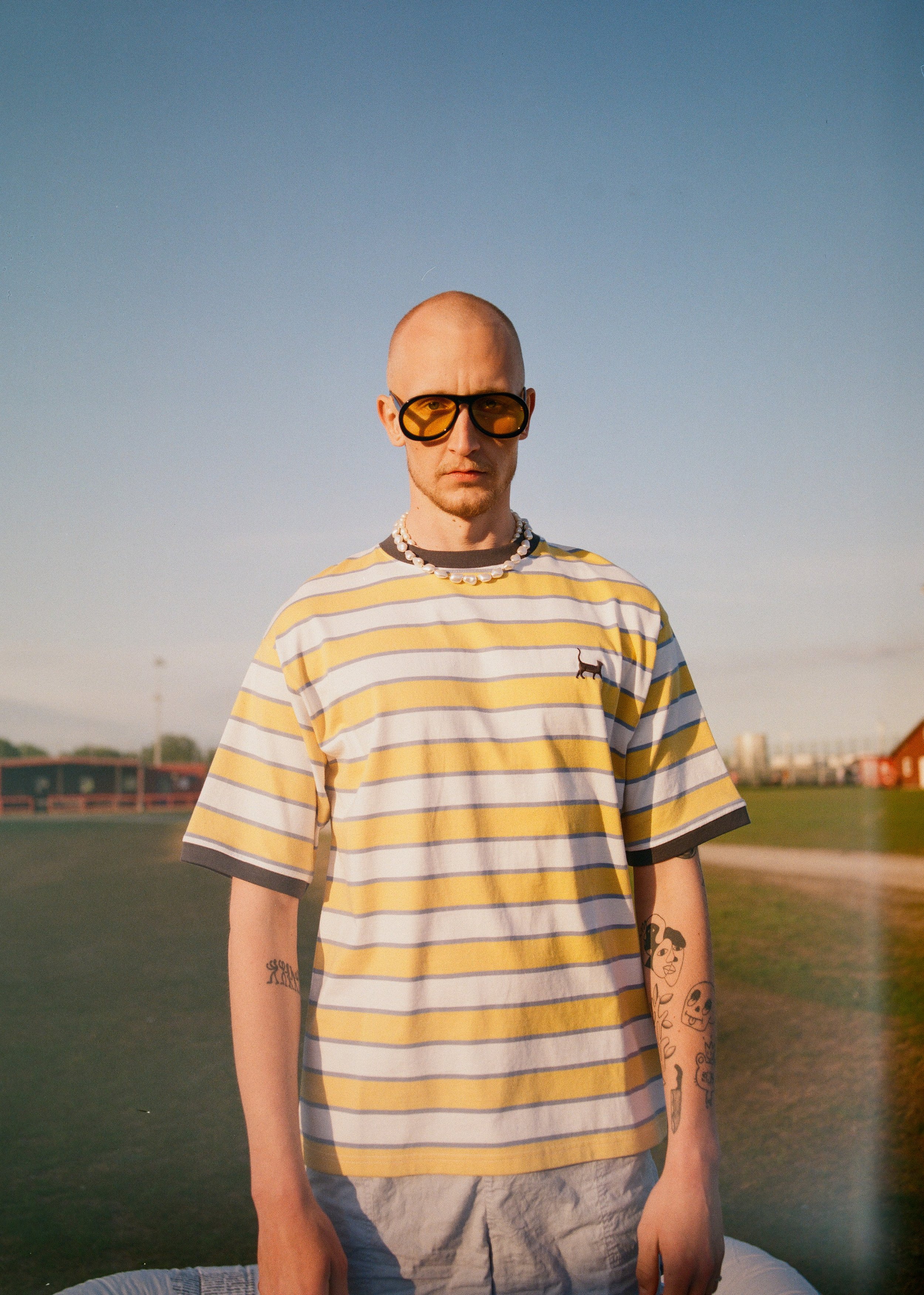 A young man with a shaved head wearing large yellow sunglasses, a pearl necklace, a striped yellow and white t-shirt with a small black dog logo, and tattoos on his arms, standing outdoors during golden hour.