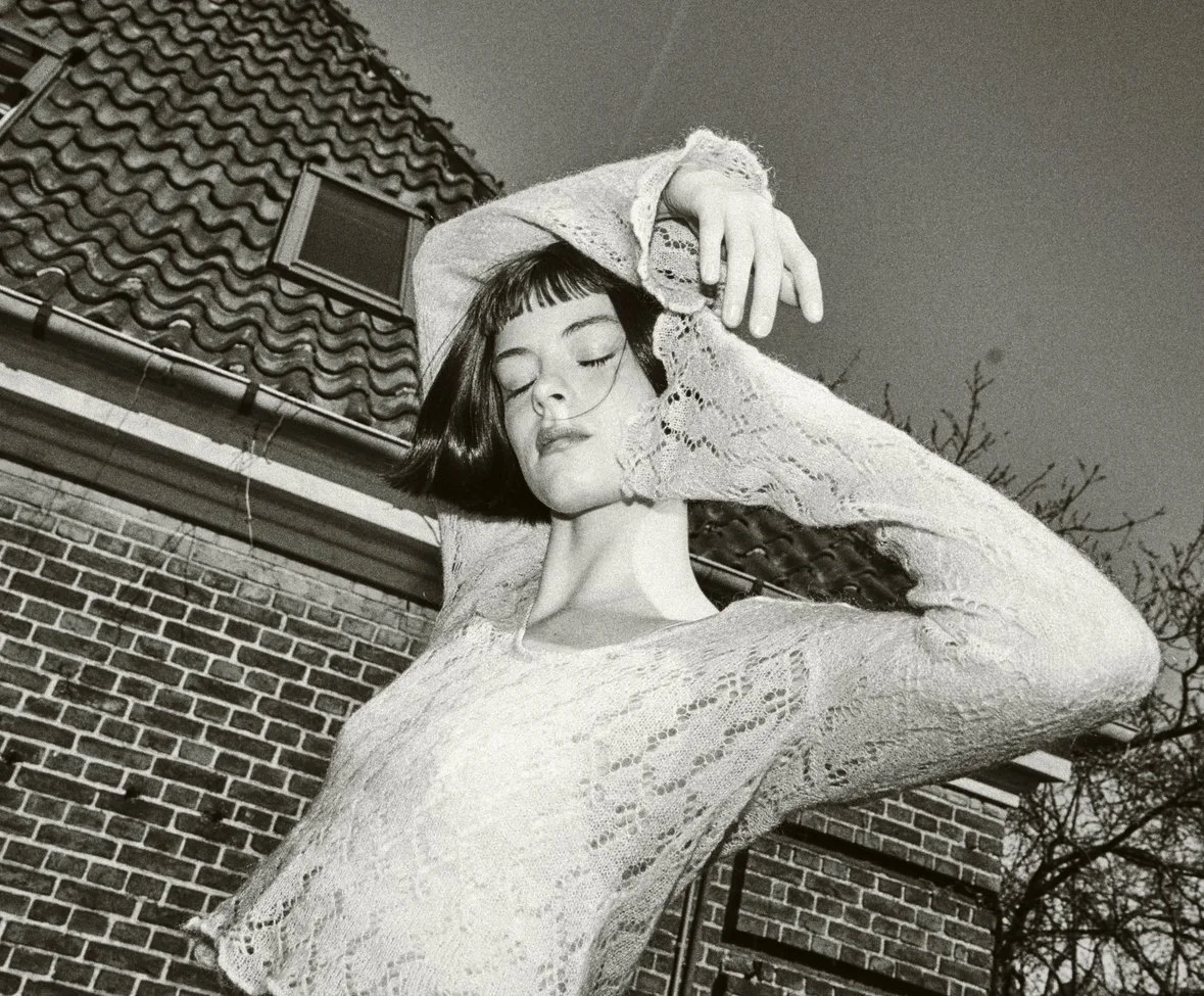 Black and white photo of a woman with short dark hair and bangs, eyes closed, wearing a lace top, with her arms raised above her head. She is outside near a brick building with a tiled roof and a small window.