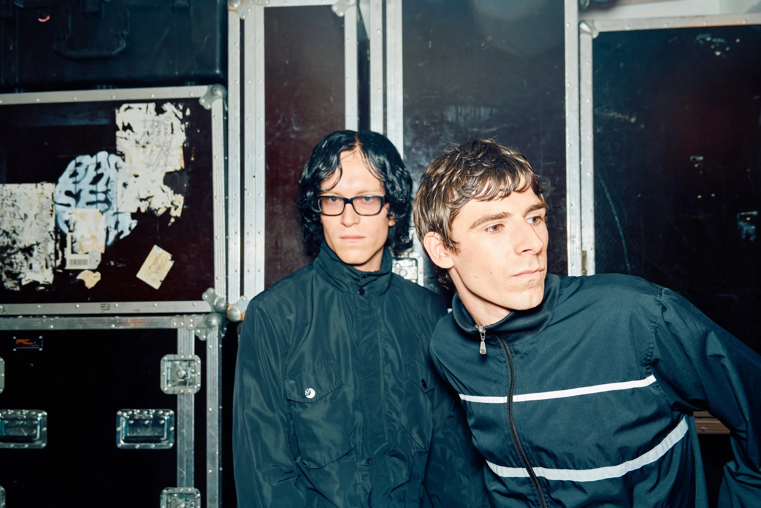 Two young men in black windbreakers, one with glasses and curly black hair, standing against a dark, industrial background with metal crates and torn posters.