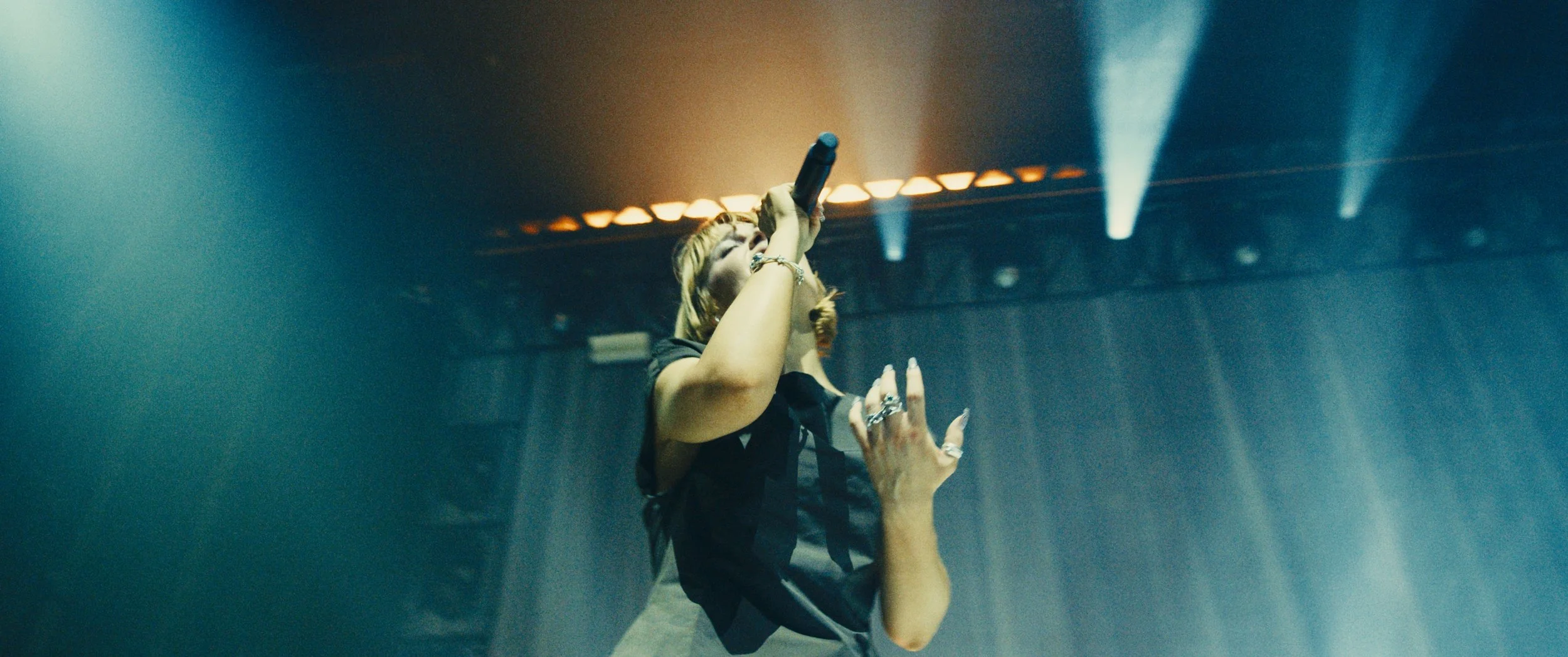 A woman singing into a microphone on stage, illuminated by stage lights, with a hand reaching up near her.