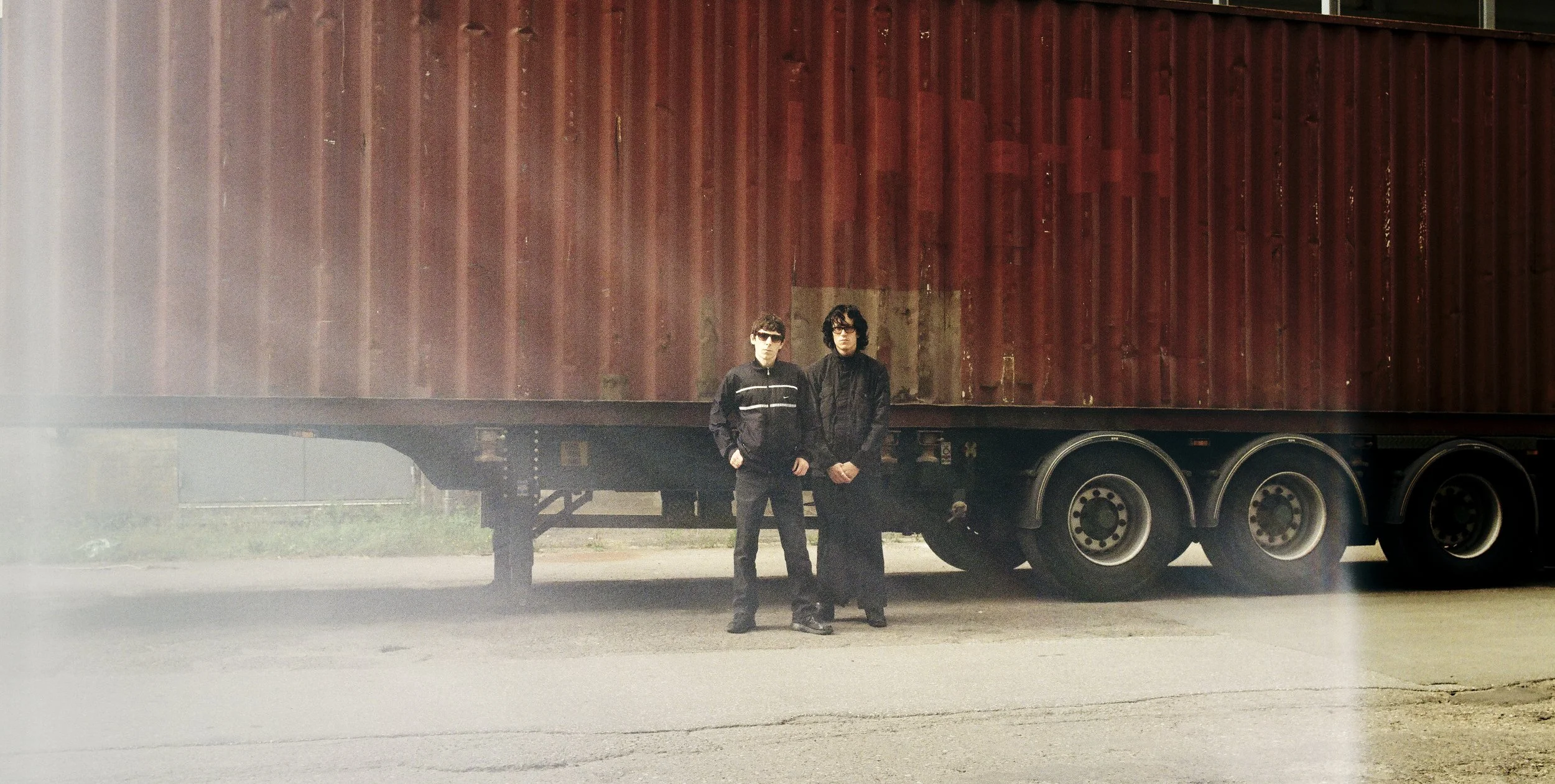 Two men in dark clothing and sunglasses standing in front of a large red shipping container on a truck trailer.