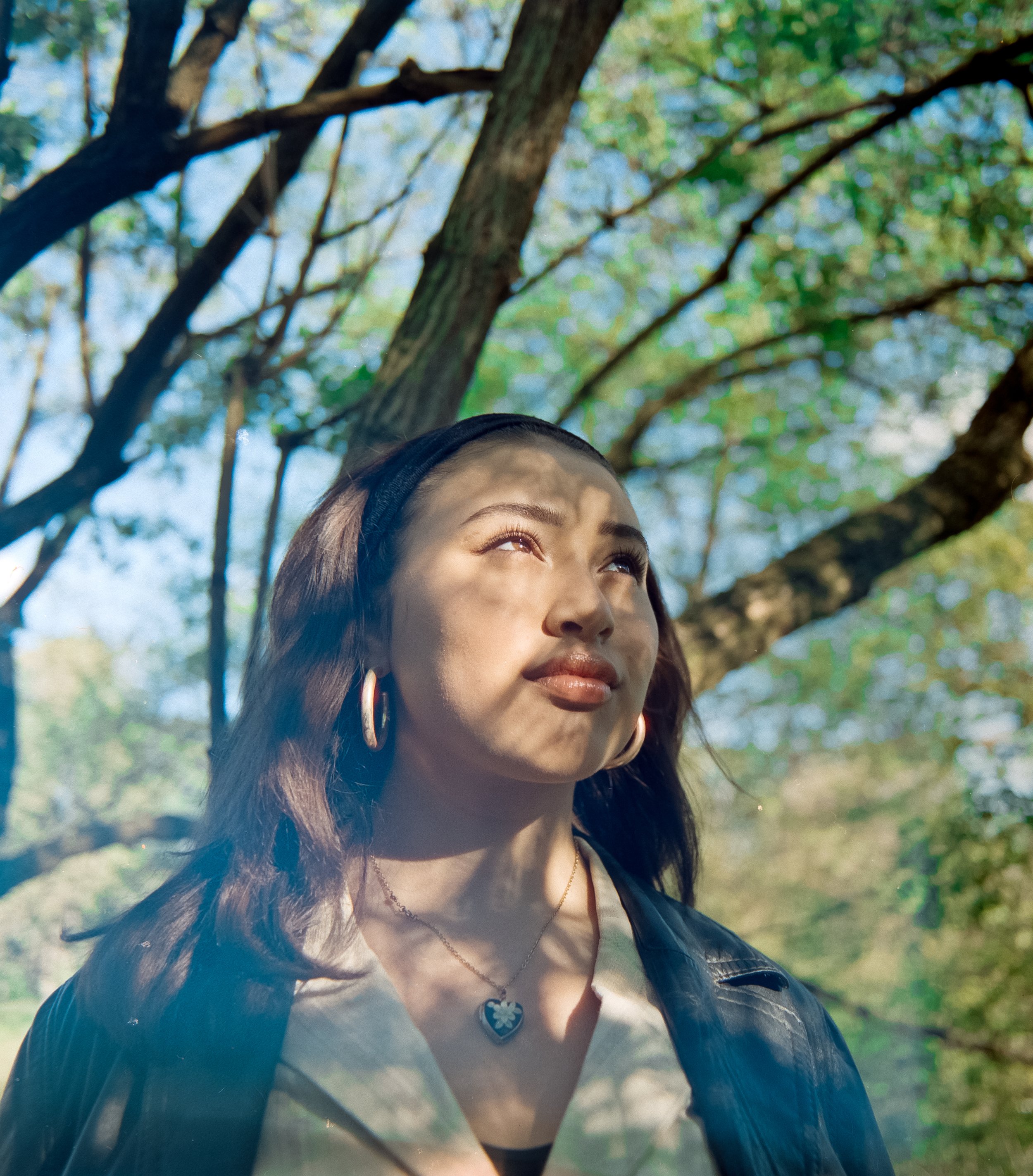 A woman with long wavy hair, wearing hoop earrings, a heart-shaped pendant necklace, a light-colored blouse, and a black headband, standing outdoors under trees with sunlight filtering through the leaves.