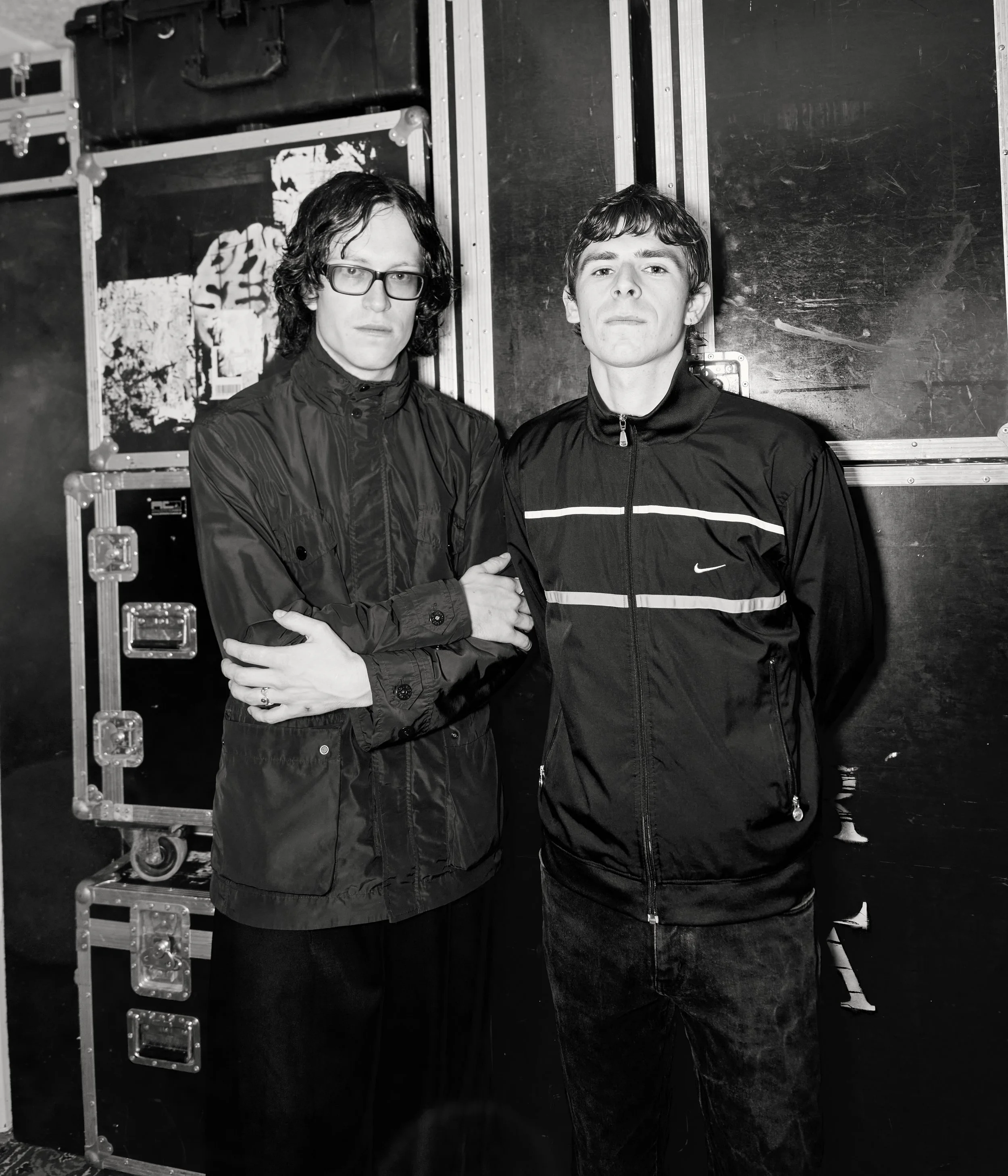 Two men standing next to each other against a background of equipment cases, with serious expressions, one wearing glasses and the other in a Nike sports jacket.