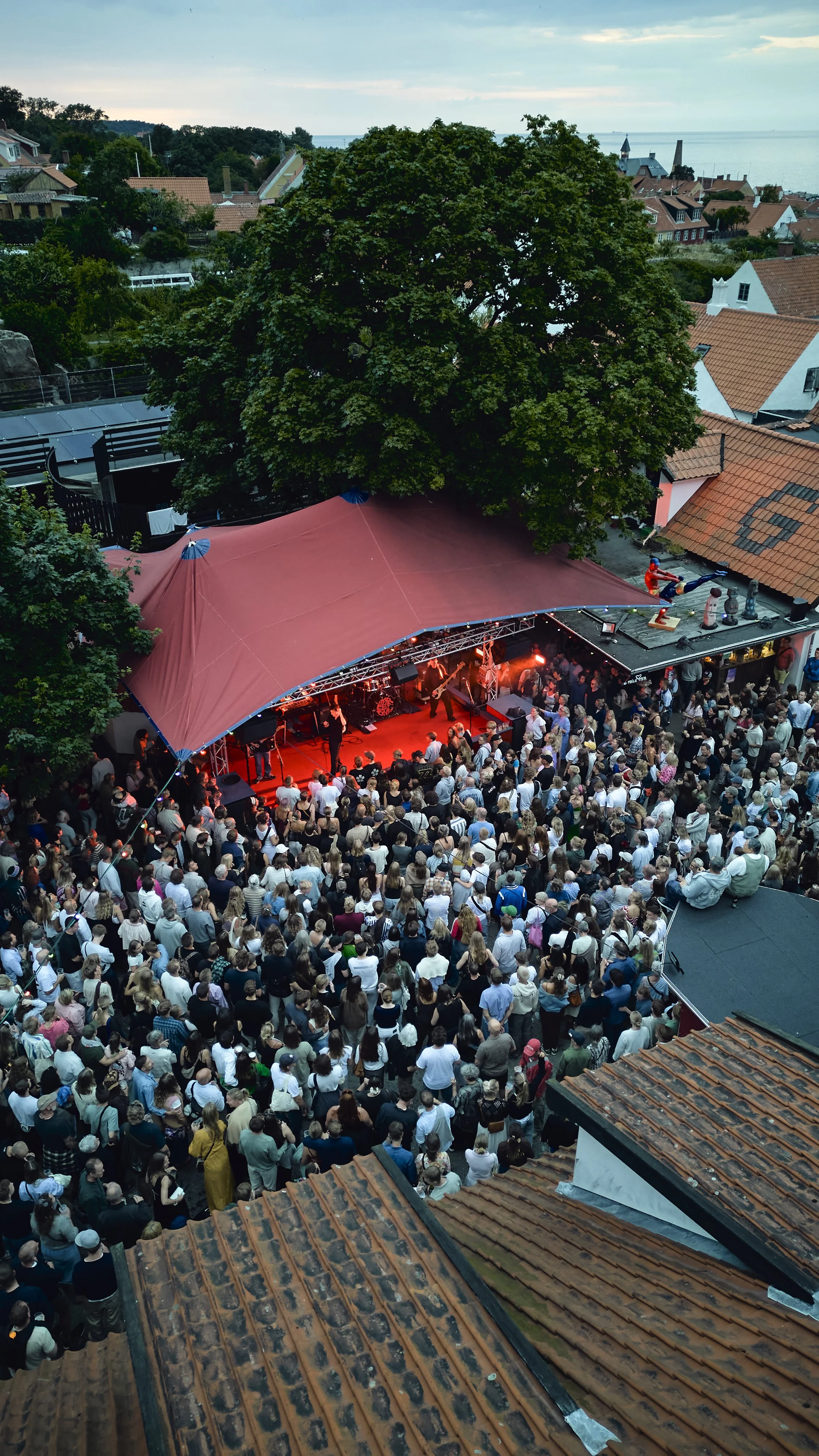 Overhead view of a large crowd gathered under a canopy at an outdoor concert, with a band performing on stage. The setting is amidst a neighborhood with houses and trees, overlooking the ocean in the background.