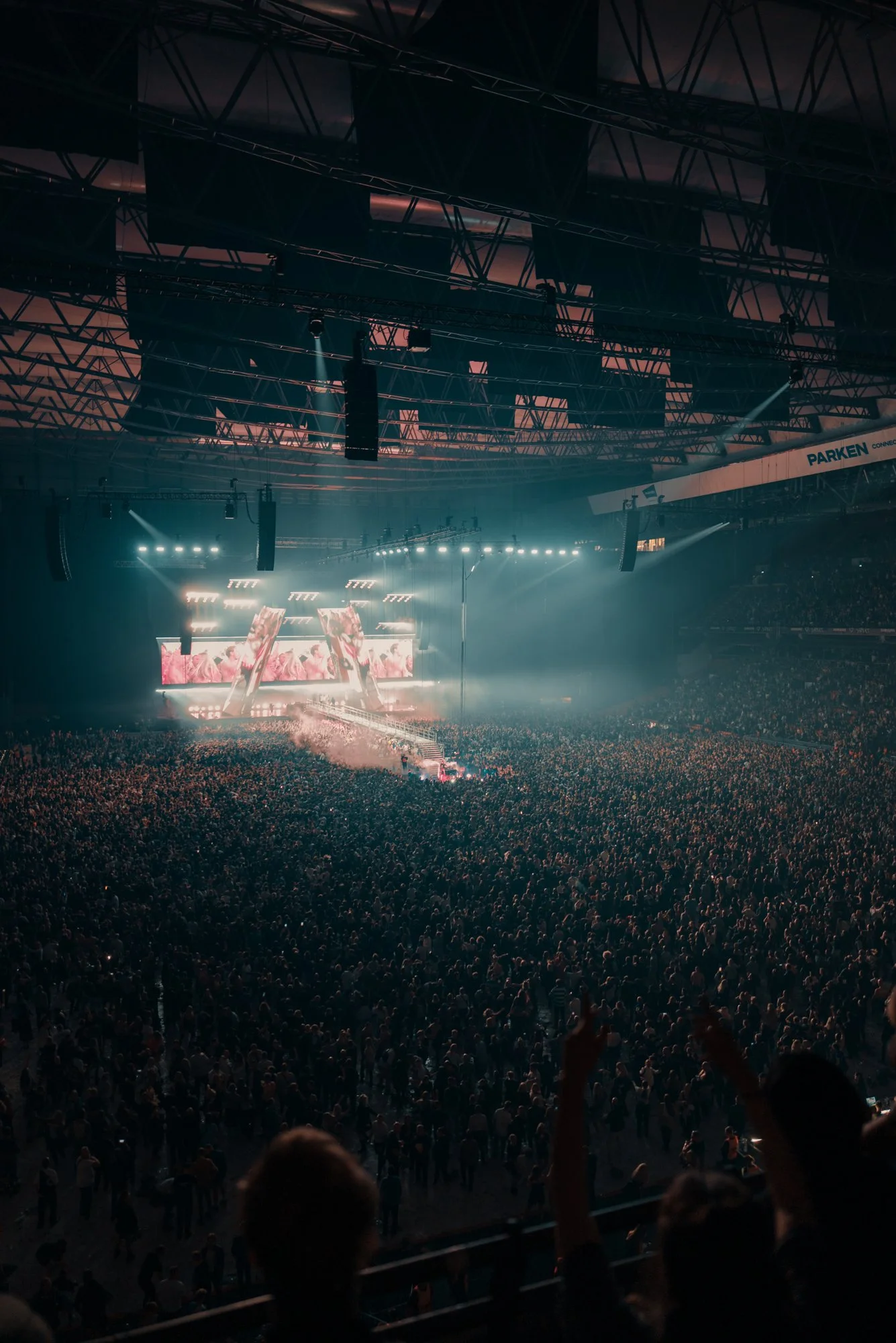 Large crowd attending a concert or music event inside an indoor arena with stage lighting and screens.