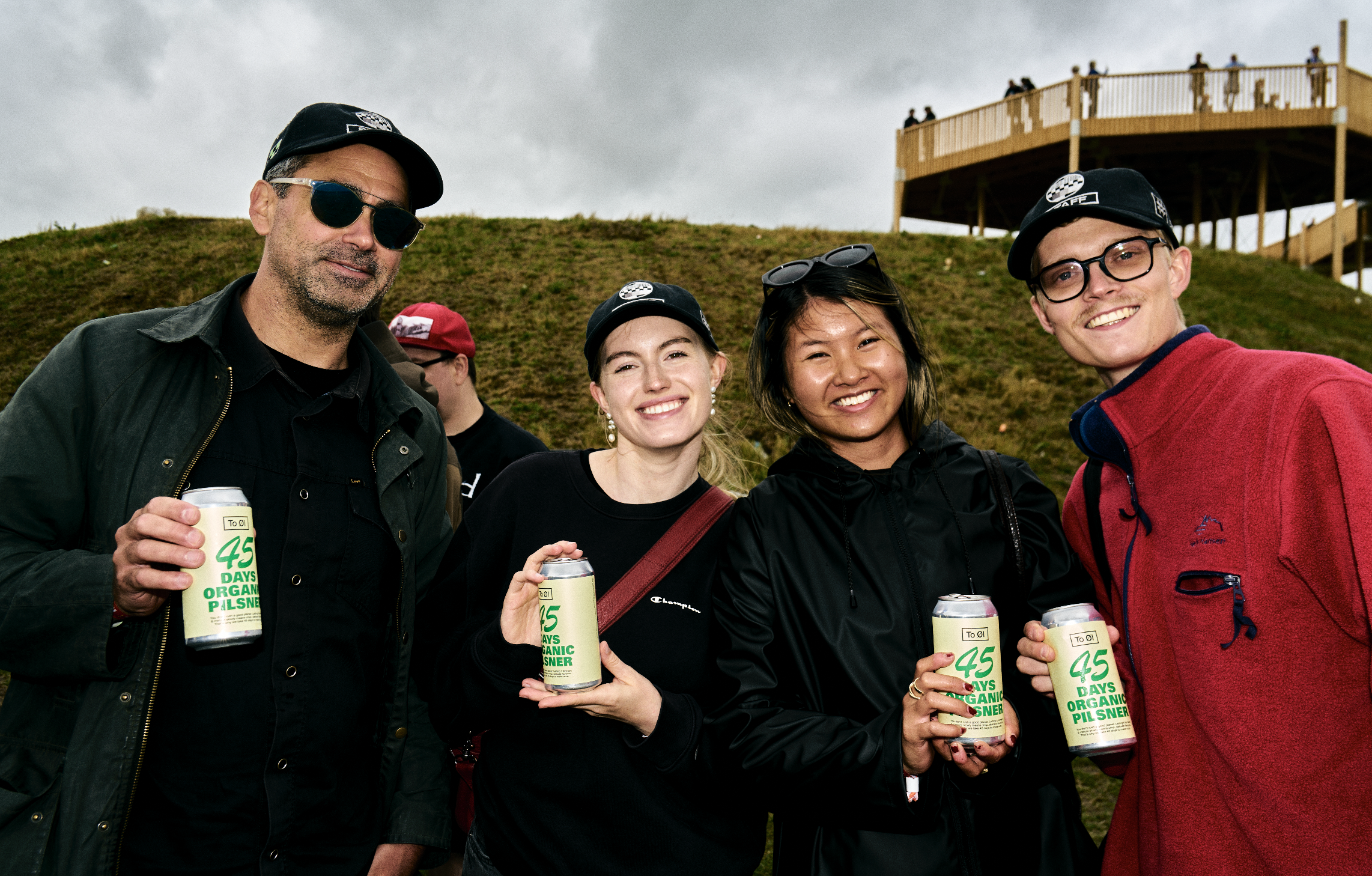 Group of four friends smiling outdoors, holding cans of organic pilsner beer, standing on a grassy hill with a wooden observation deck in the background on overcast day.