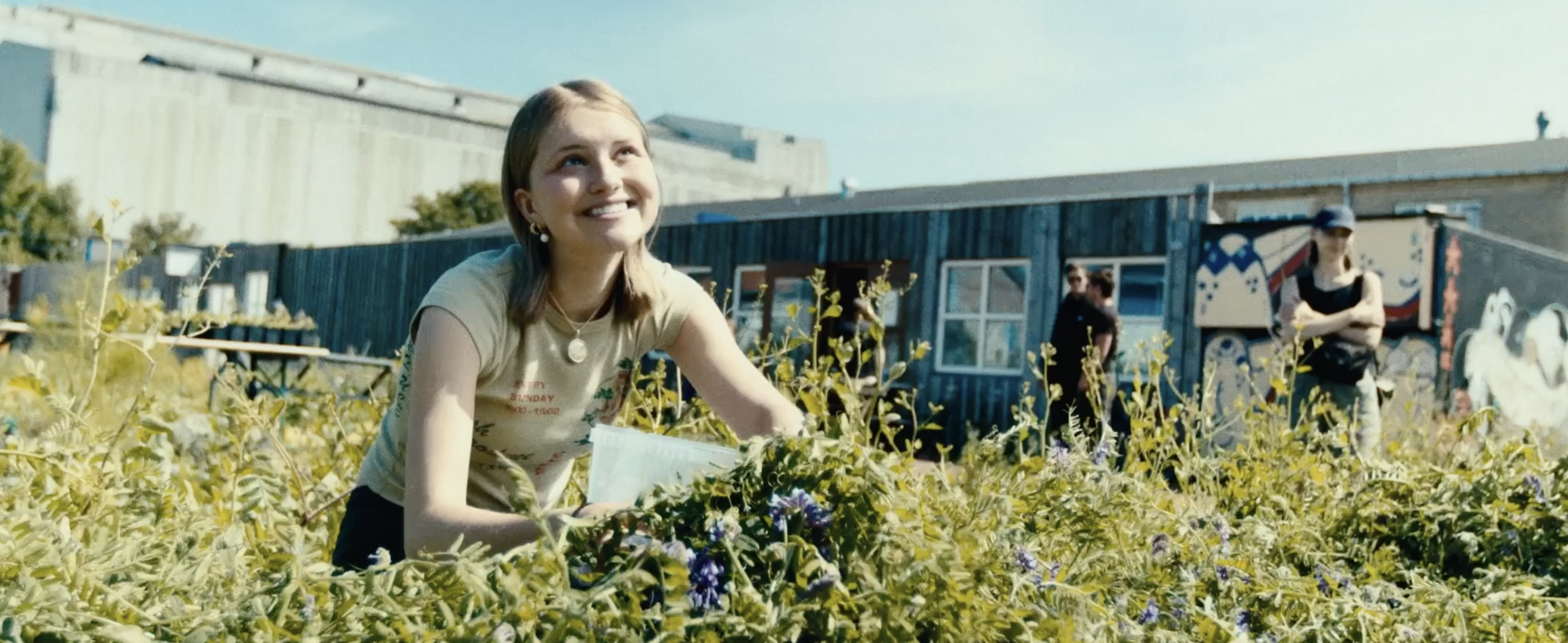 A smiling young woman with shoulder-length blonde hair, wearing a beige t-shirt and jewelry, is crouching among yellow flowering plants outdoors, with a blue building and other people in the background.