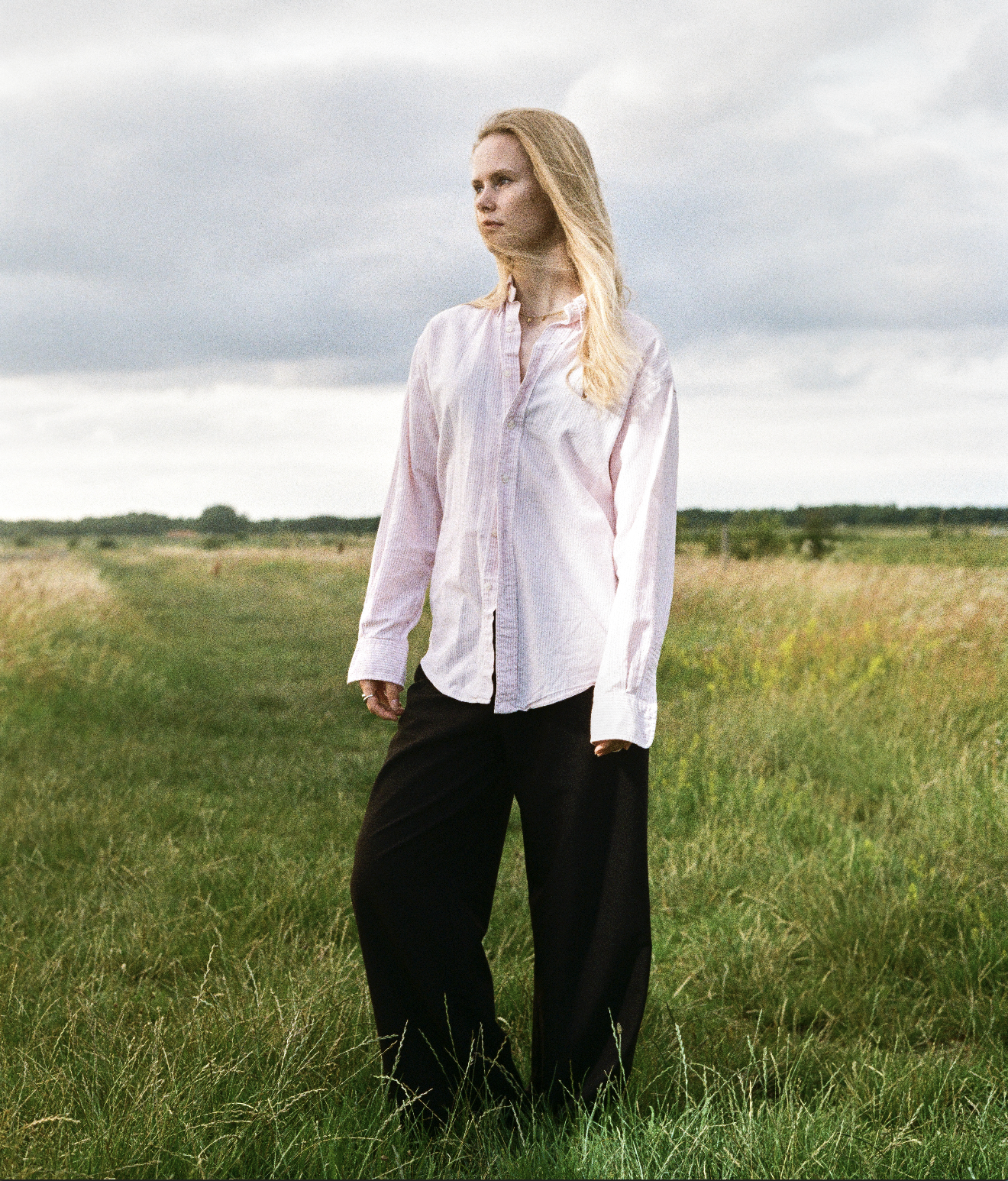 A woman with blonde hair standing in a grassy field under a cloudy sky, wearing a light pink button-up shirt and black wide-leg pants.