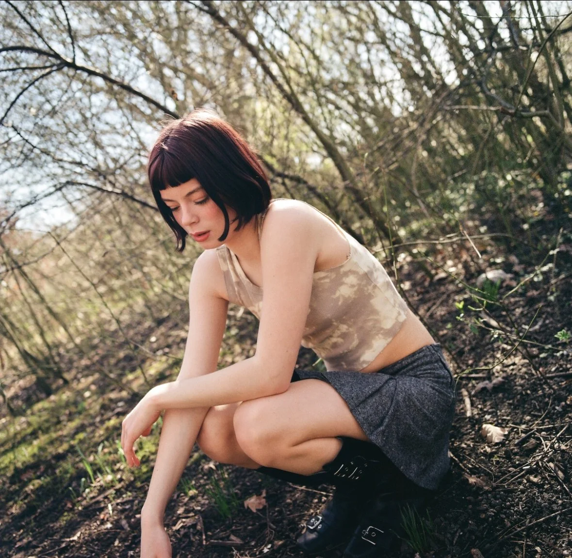A young woman with dark hair and bangs sits on the ground in a wooded area, looking down thoughtfully surrounded by trees and sunlight.