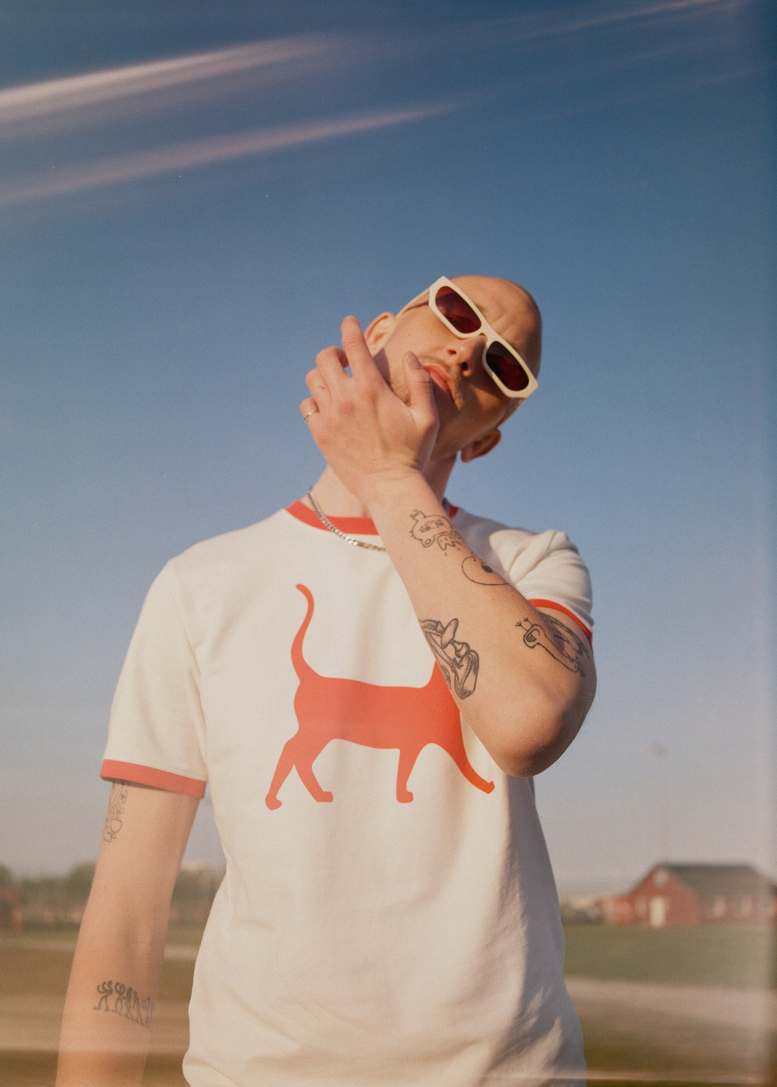 A person wearing sunglasses and a white T-shirt with an orange cat graphic, standing outdoors against a blue sky with a few clouds.