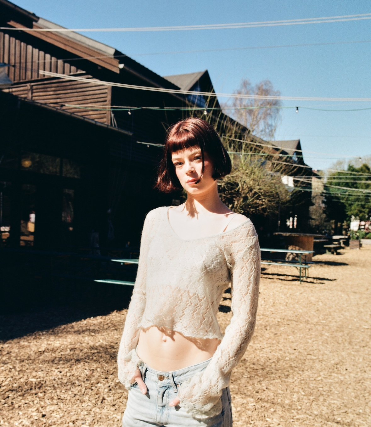 A young woman with short dark hair standing outdoors on a sunny day, wearing a cream-colored lace long-sleeve top and light blue jeans.
