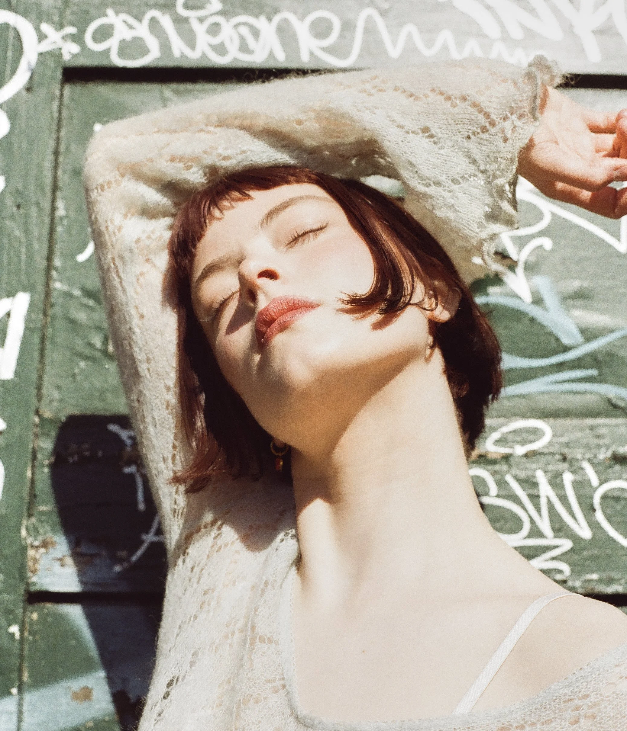 A woman with short brown hair and closed eyes relaxing outdoors on a sunny day, lying on a graffiti-covered wooden surface with her arm above her head, wearing a white lace top.