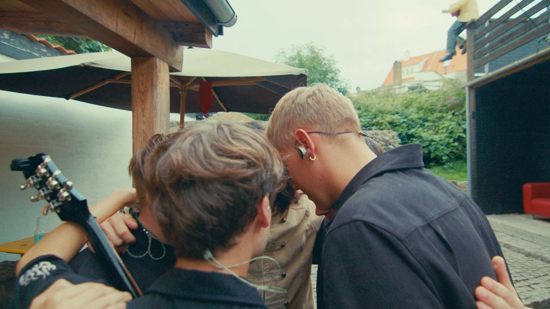 Four people standing close together outdoors with their heads bowed and arms around each other, possibly in a hug or prayer, with umbrellas and greenery in the background.