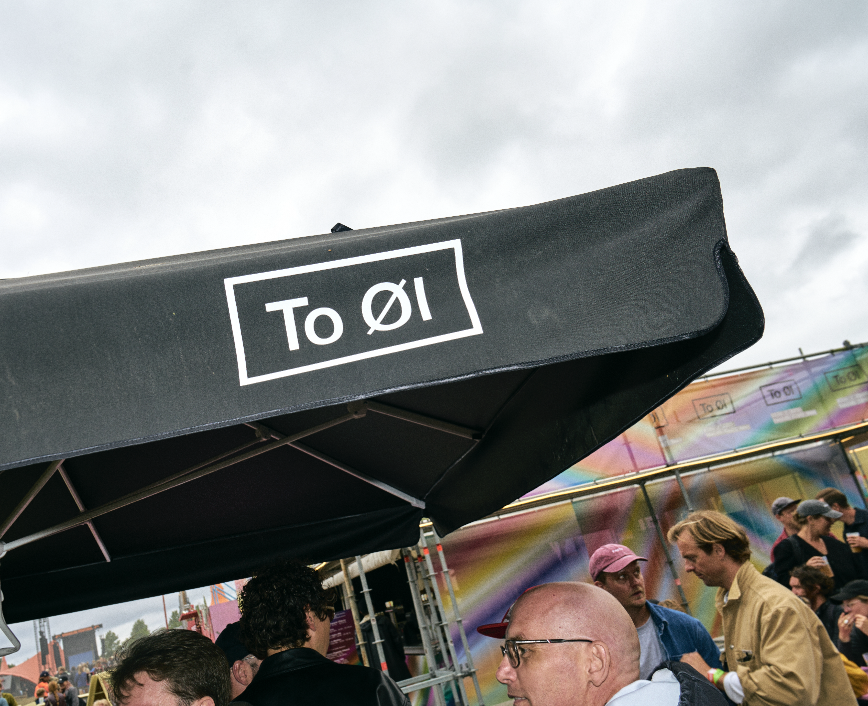 Crowd of people at an outdoor event or festival. A black tent with a sign reading 'To Øl' is visible, with a colorful background and overcast sky.