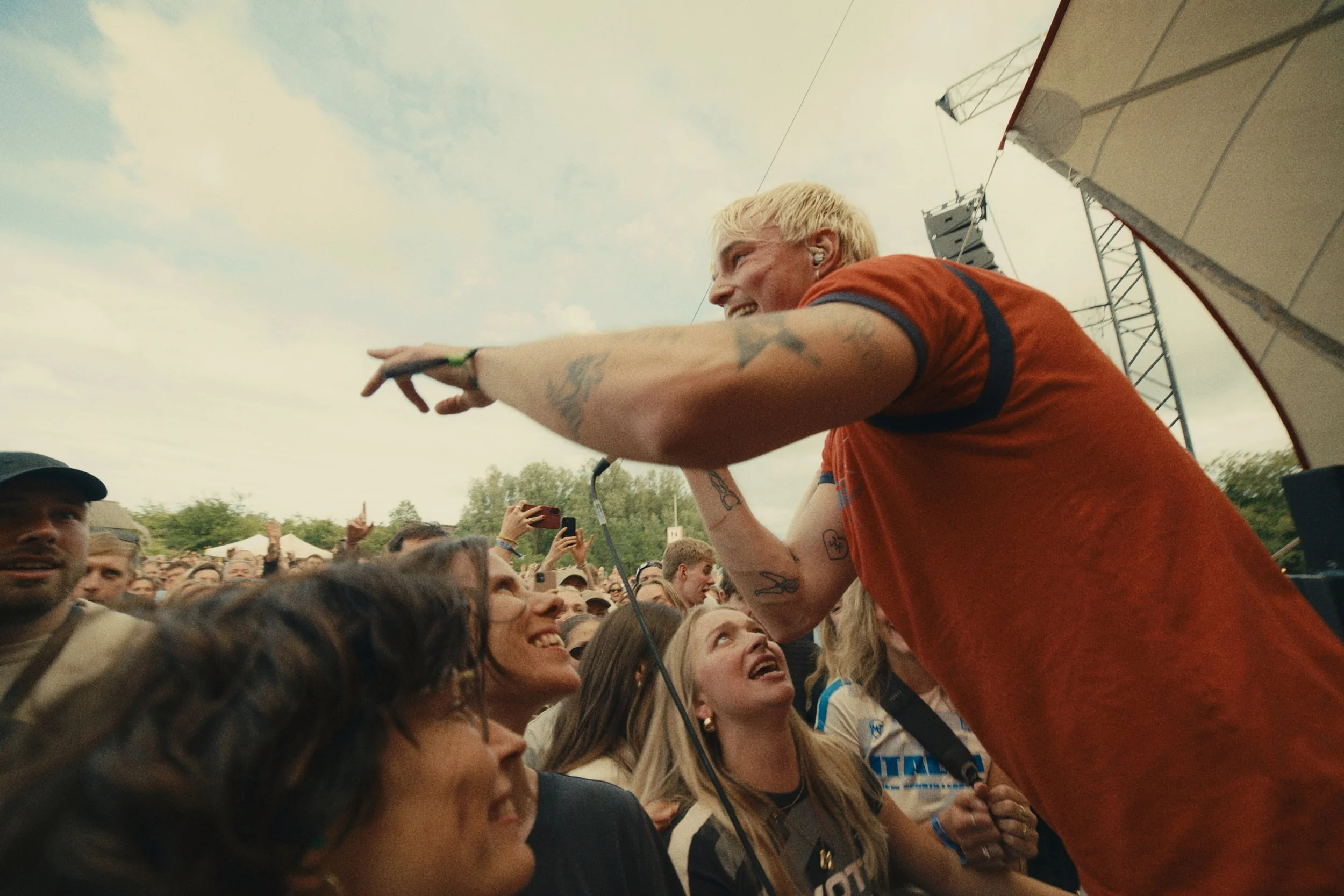 A man with blonde hair, tattoos on his arm, wearing an orange shirt, is leaning over a crowd of people, smiling, as he interacts with them at an outdoor event. Several people in the crowd are looking up and taking photos, with a partly cloudy sky and