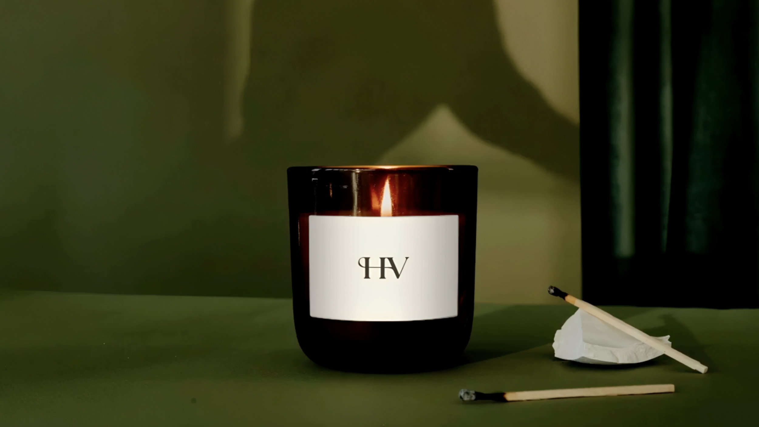 Tänt doftljus i brunt glas med Hotell Villans monogram HV på etiketten, mot en mörkgrön bakgrund med tändstickor bredvid.