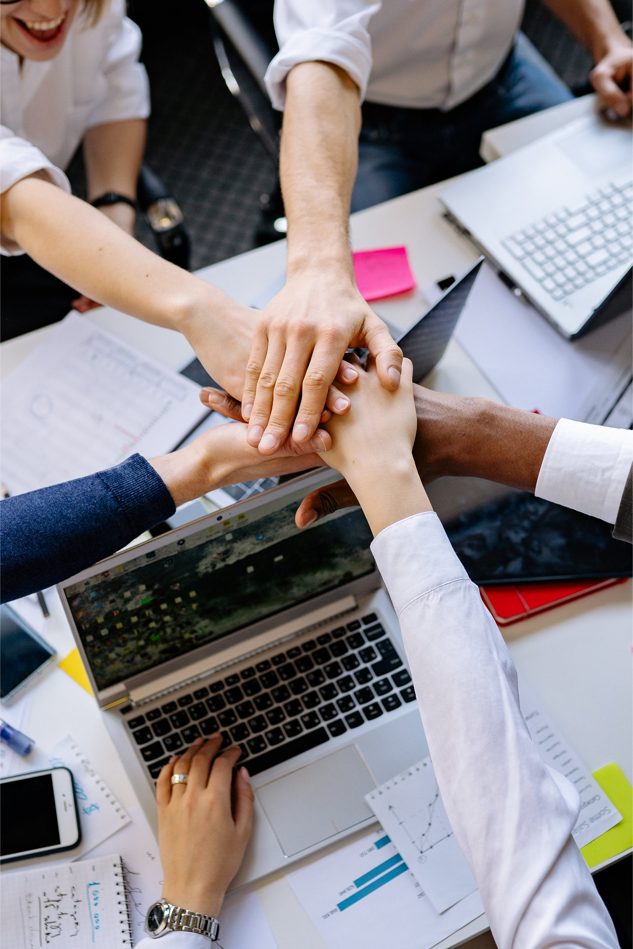 A group of people in a business meeting joining their hands together over an office table with laptops, notebooks, and documents.