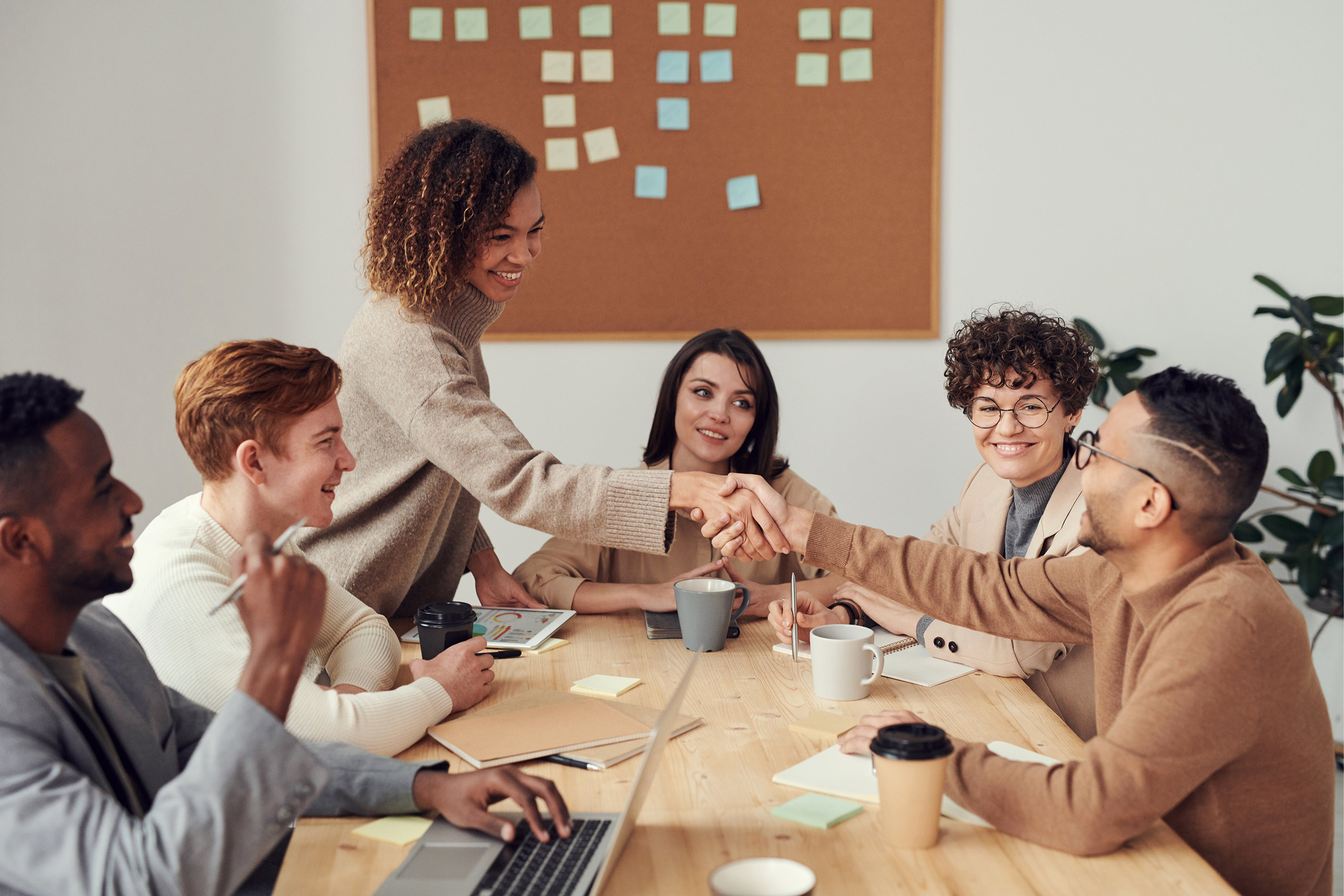 Group of six diverse young professionals in a meeting room, smiling, shaking hands, and having a discussion around a wooden table with laptops, notebooks, cups, and sticky notes.