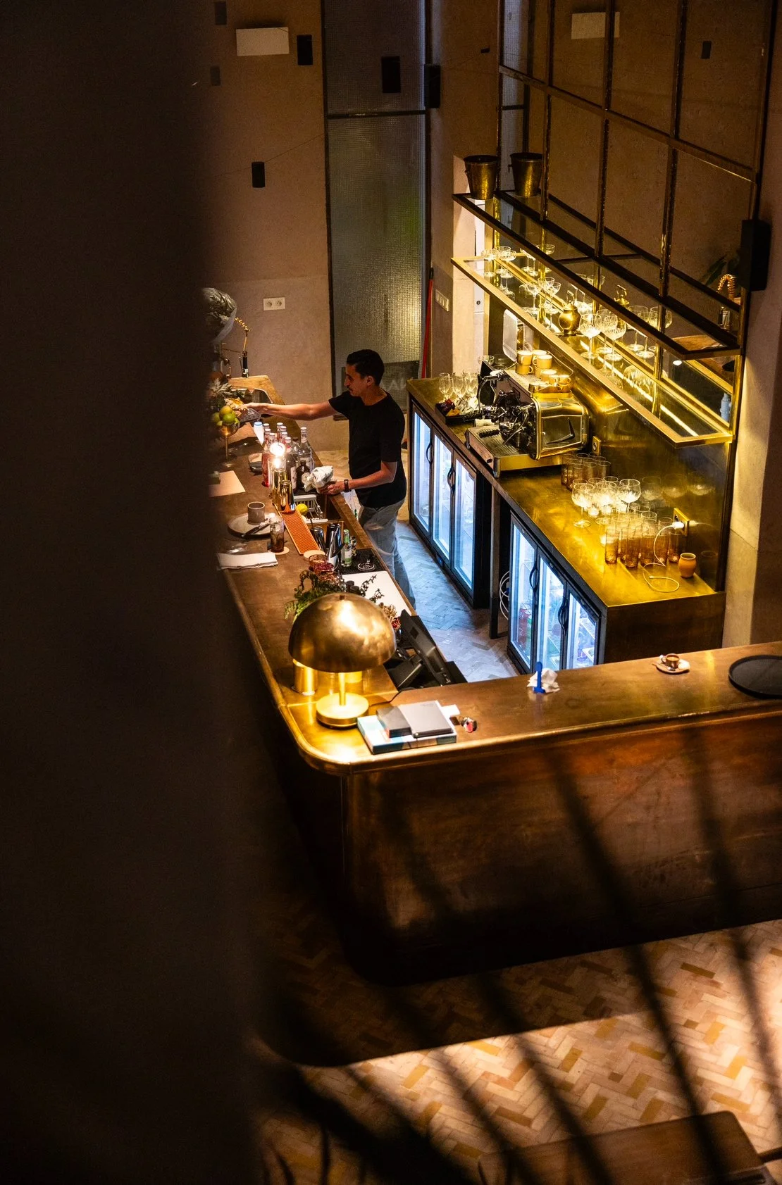 Naama Marrakech. Overhead view of a stylish bar with a bartender working, featuring a wooden counter, large golden lamps, glassware, and drink refrigerators.