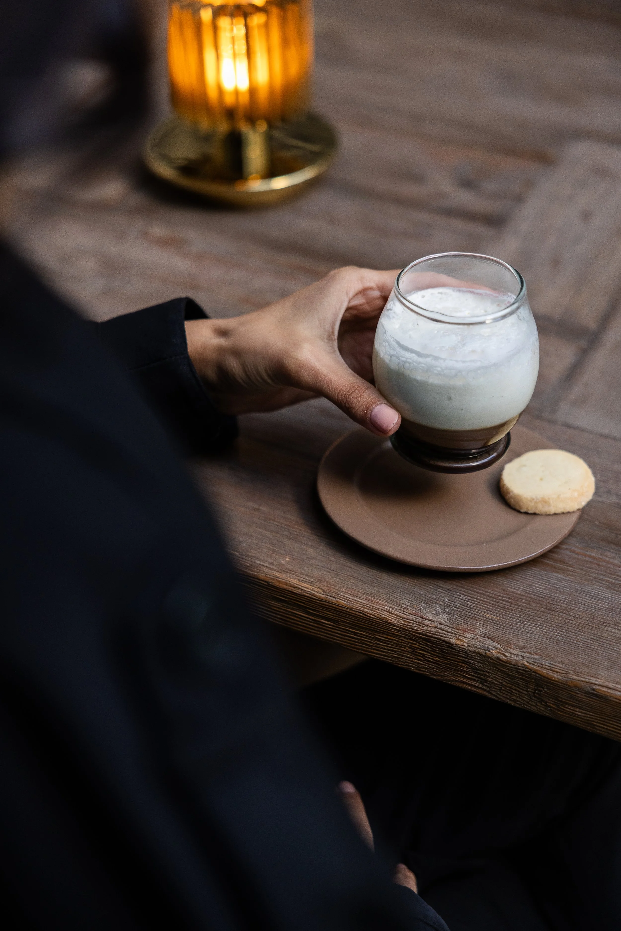 Naam. Person holding a glass of frothed milk on a wooden table next to a plate with a cookie and a lit candle in the background.