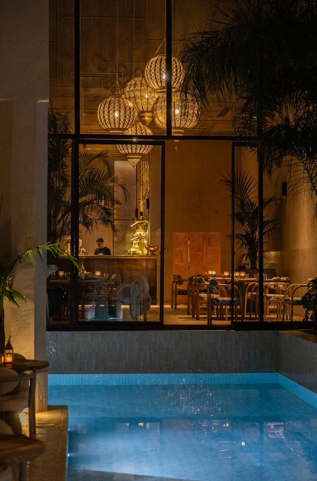 Naama Restaurant interior with hanging lamps, tropical plants, and swimming pool view at night