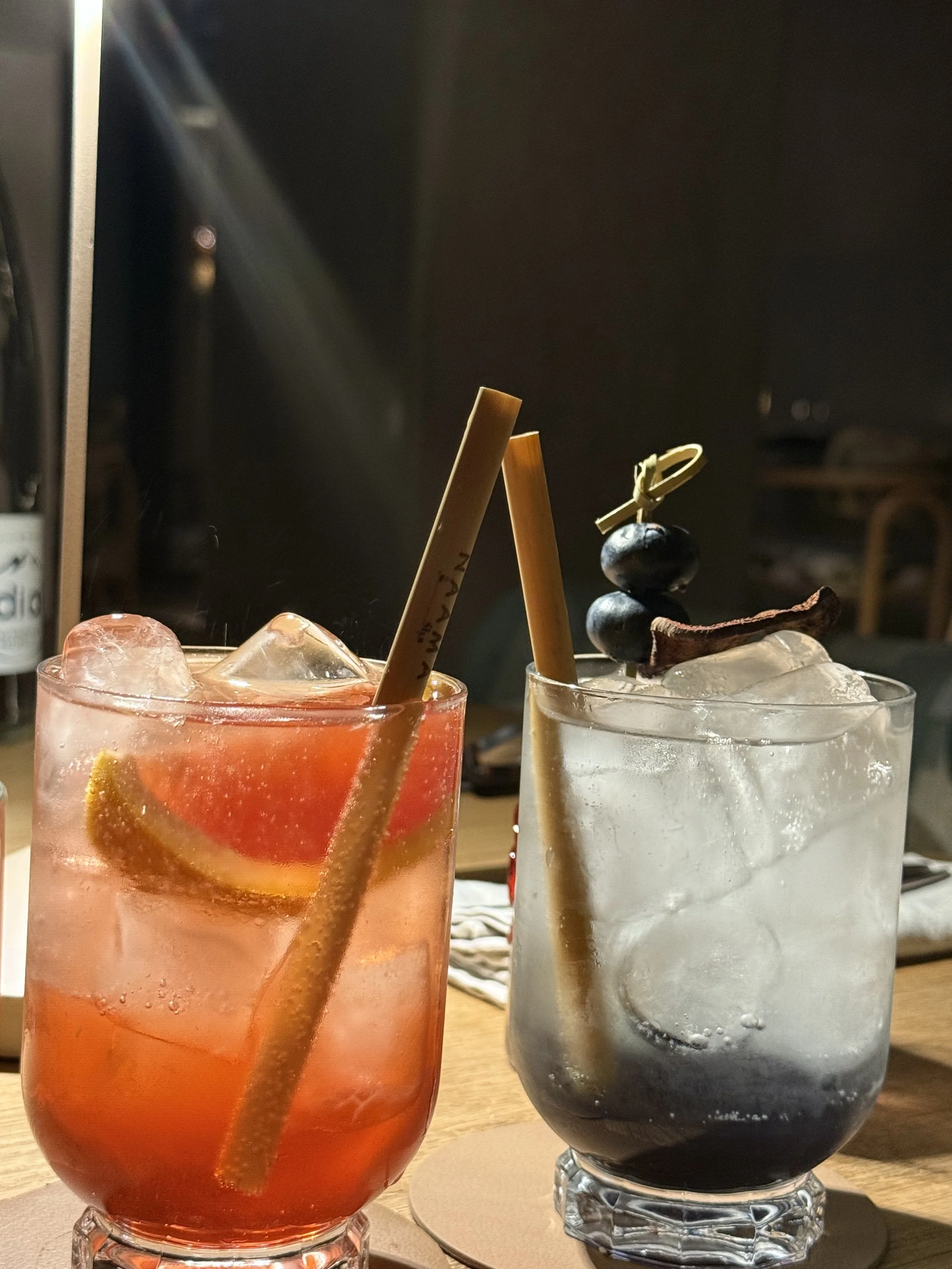 Two cocktails with ice, lemon wedge, and straw on a wooden table, one red and one clear with dark berries and a strip of bacon garnish.