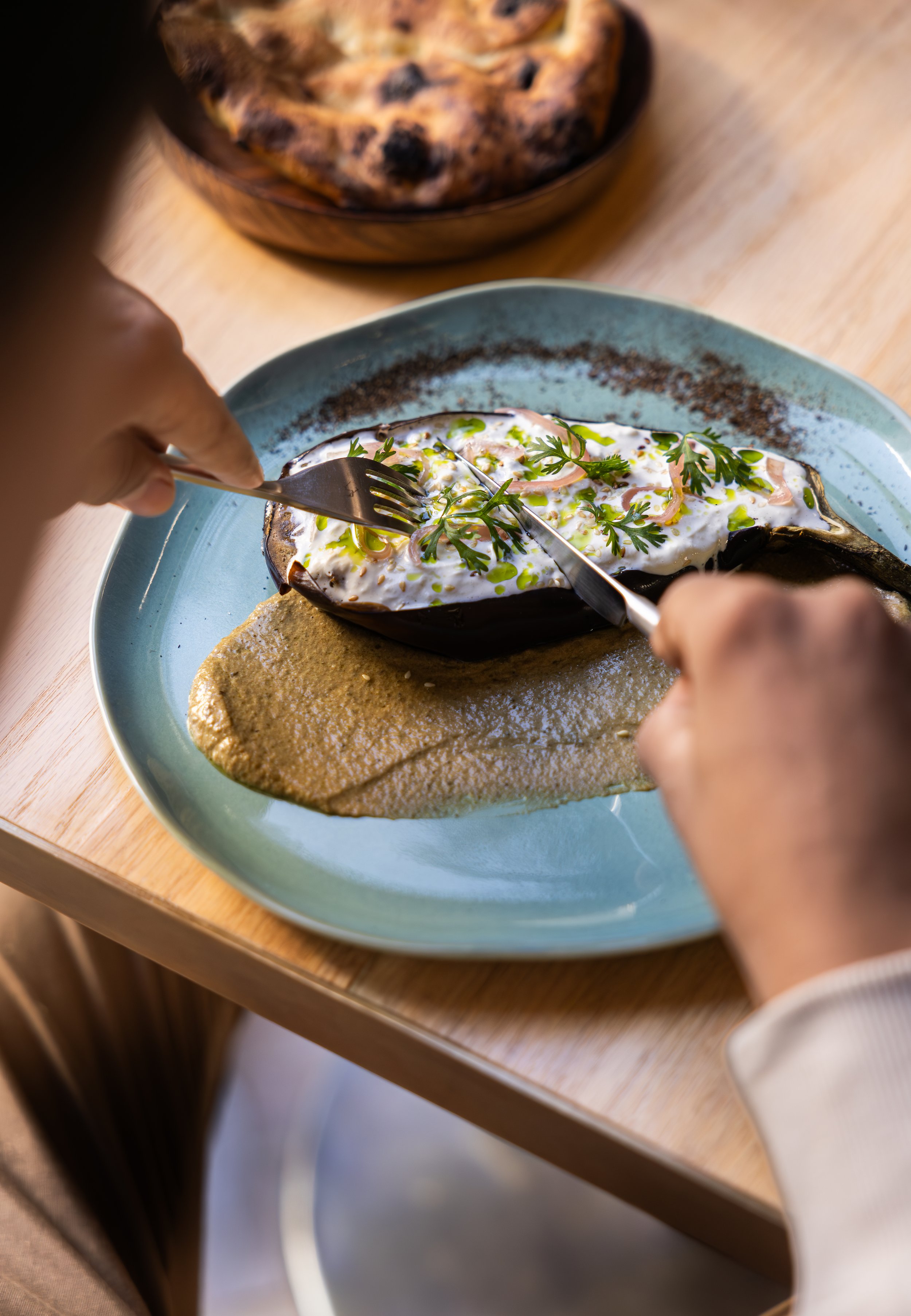 A person using a fork and knife to eat a baked eggplant topped with creamy sauce and herbs on a blue plate, with a bread dish in the background.